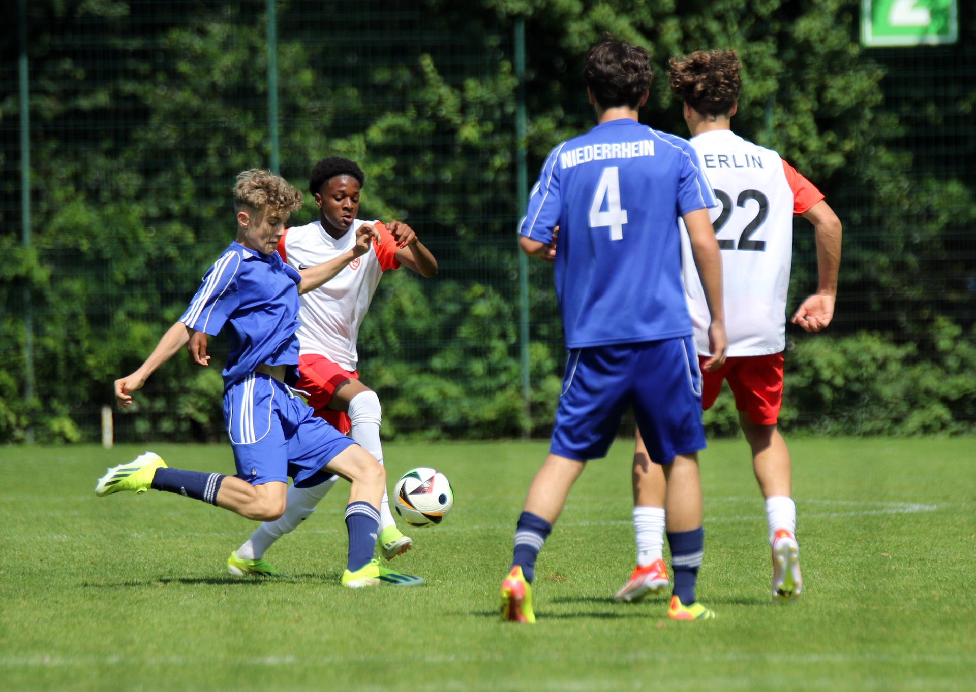 [{"clang_id":1,"value":"Fußballspiel: Junge mit blonden Haaren schießt auf das Tor, verfolgt von Gegenspielern in blau-weißem und rot-weißem Trikot auf einem grünen Rasenfeld."}]
