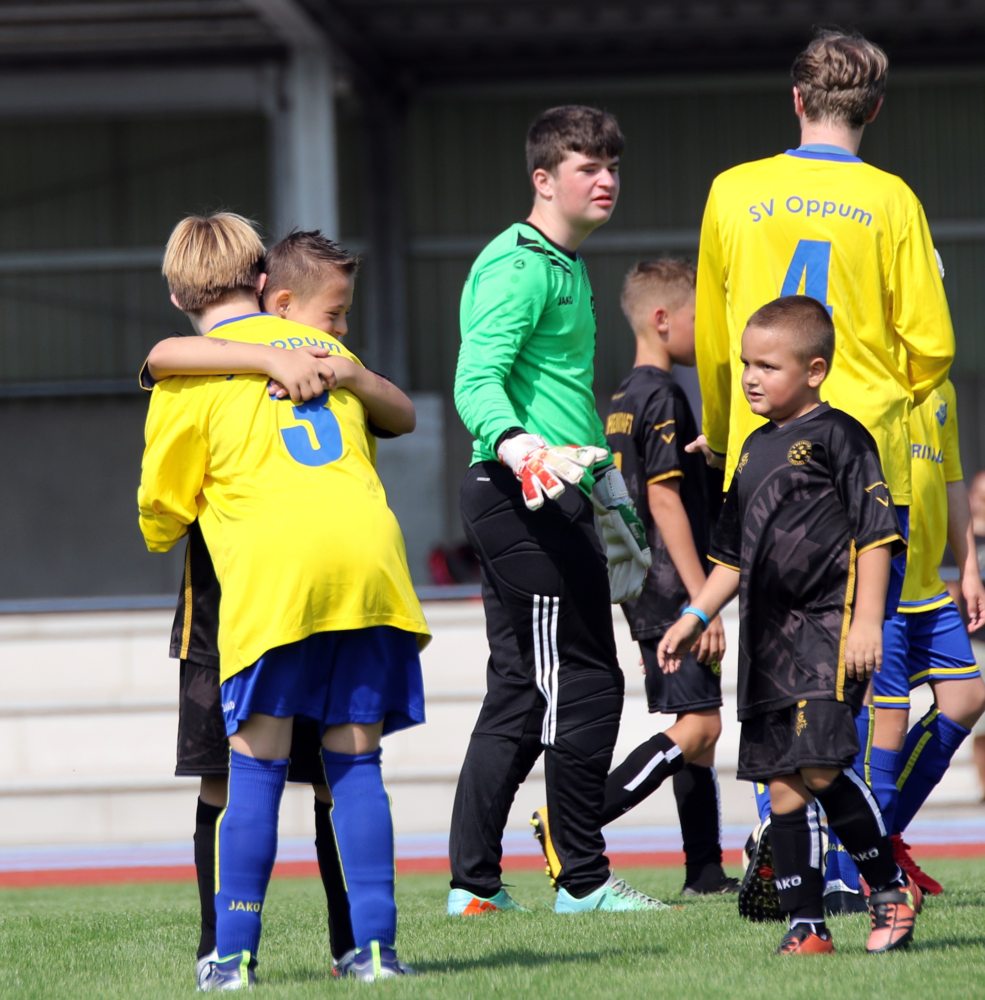 [{"clang_id":1,"value":"Fußballspieler, Kinder und ein Jugendlicher im Fußballoutfit stehen auf einem Rasenplatz. Zwei Kinder umarmen sich. Ein Jugendlicher, vermutlich Torwart, führt ein Kind an der Hand."}]