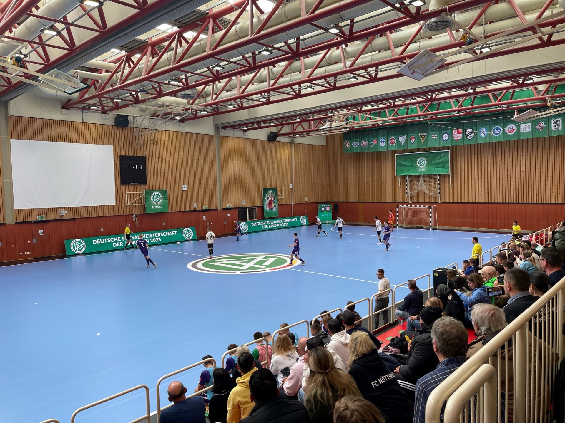[{"clang_id":1,"value":"Futsal-Halle mit Spiel in Gange. Zuschauerreihen gefüllt mit Menschen. Im Hintergrund Banner mit Schriftzügen und Wappen."}]