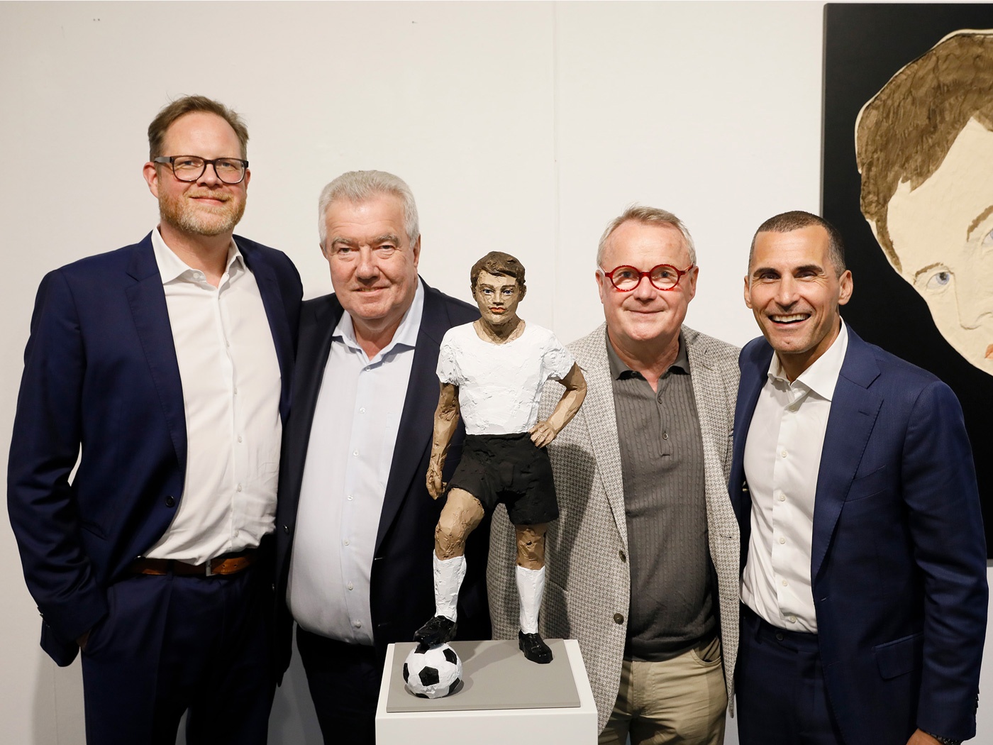 [{"clang_id":1,"value":"Vier Männer stehen vor einer weißen Wand. Zwischen ihnen eine Skulptur eines Fußballspielers auf einem Fußball. Im Hintergrund ein Porträt."}]