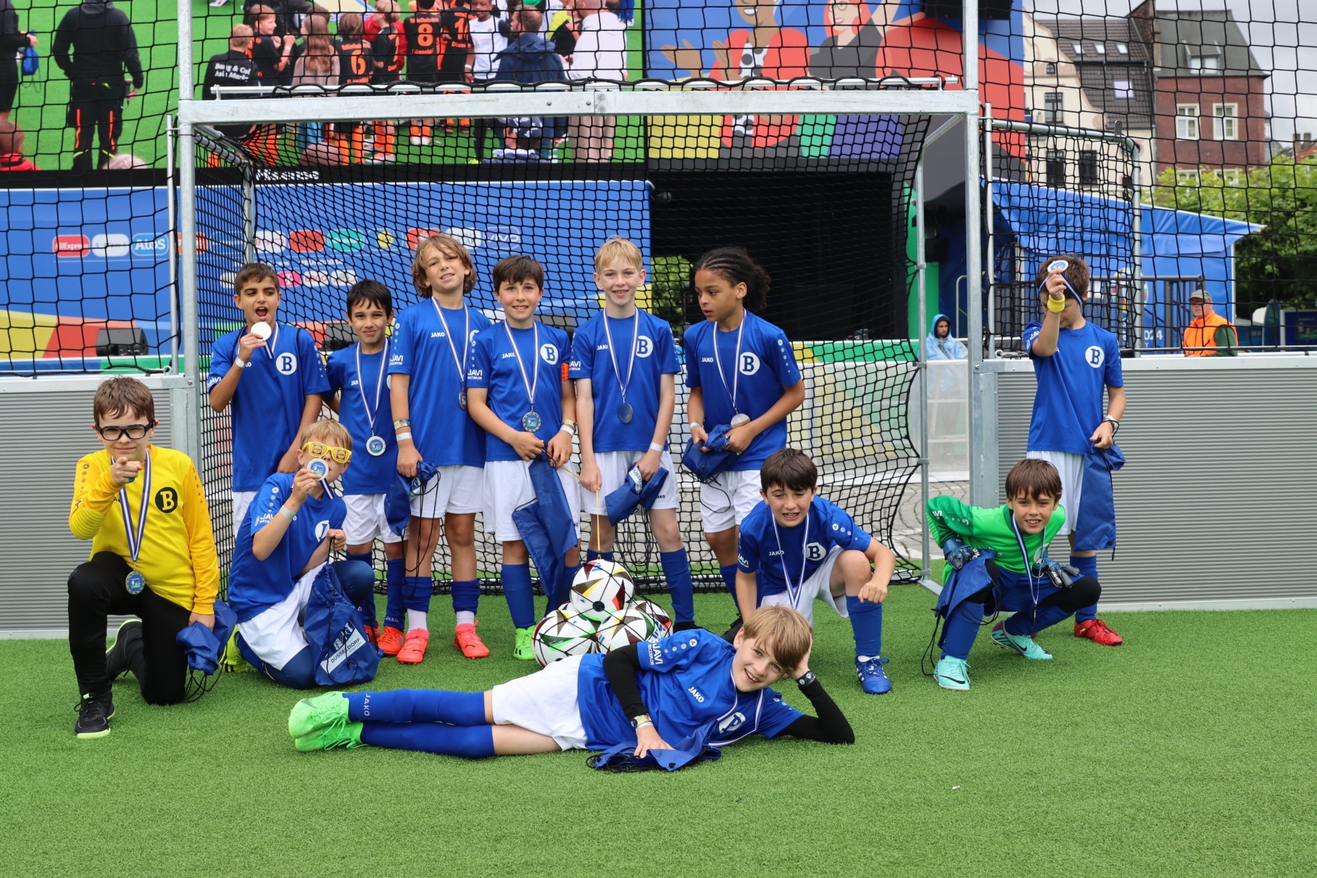 [{"clang_id":1,"value":"Fußballmannschaft mit blauen Trikots und Medaillen posiert vor einem Tor auf grünem Kunstrasen. Einige Kinder liegen oder hocken im Vordergrund."}]