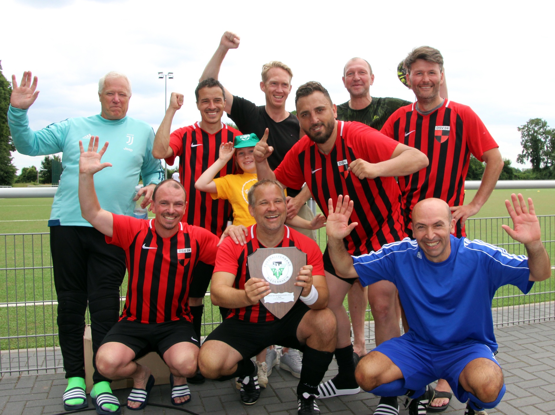 [{"clang_id":1,"value":"Fußballmannschaft in rot-schwarzen und blauen Trikots posiert mit Pokal. Mehrere Männer lächeln und winken in die Kamera, eine Frau steht mittendrin. Hintergrund: Sportplatz mit Rasen."}]