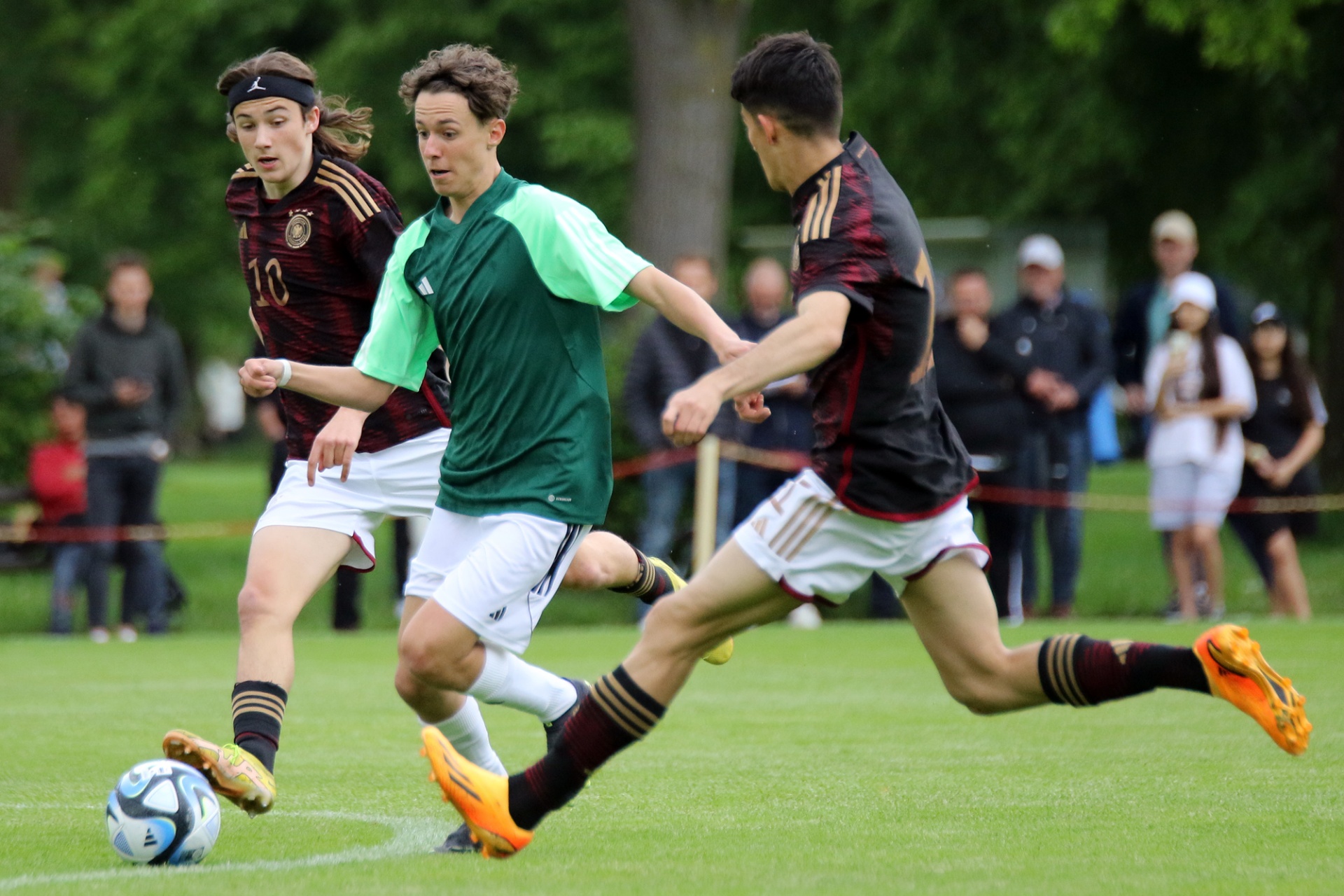 [{"clang_id":1,"value":"Drei Fußballspieler auf Grün. Ein Spieler in grün-weißem Trikot schießt auf den Ball, wird von zwei Spielern in rot-schwarzem Trikot begleitet. Hintergrund: Zuschauer und Bäume."}]