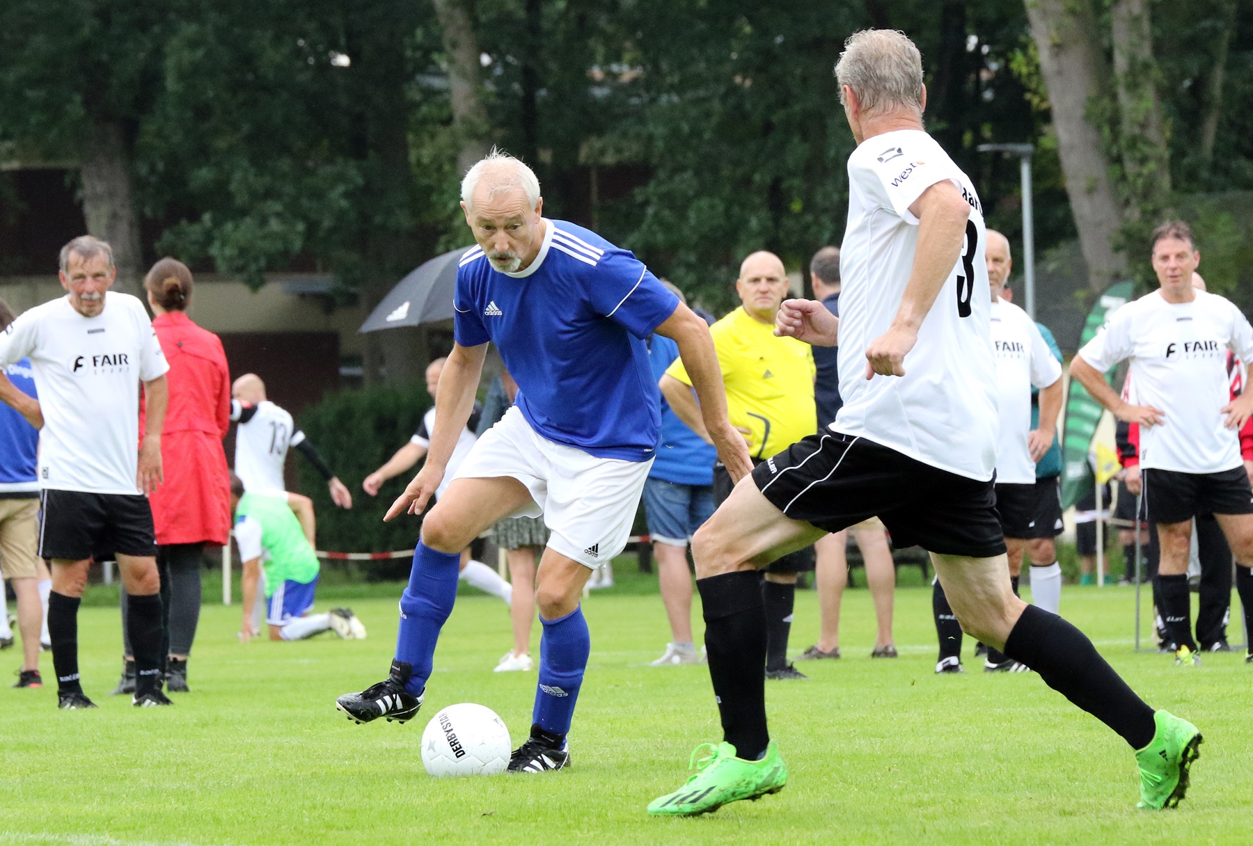 [{"clang_id":1,"value":"Zwei ältere Männer spielen Fußball auf einem grünen Rasen. Einer trägt blaues Trikot, der andere weiß-schwarzes. Im Hintergrund weitere Personen und Bäume."}]