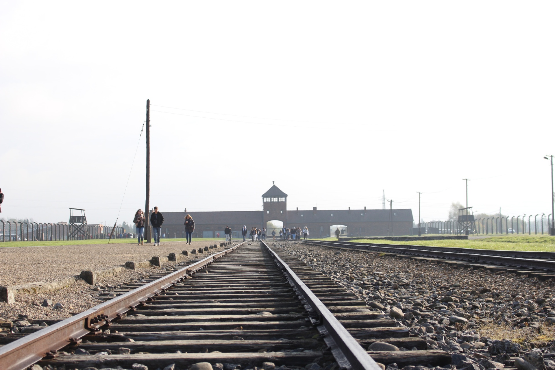 [{"clang_id":1,"value":"Eisenbahnschienen führen auf ein Gebäude im Hintergrund, vermutlich ein historischer Ort. Menschengruppen sind auf einem Weg neben den Schienen und vor dem Gebäude zu sehen."}]