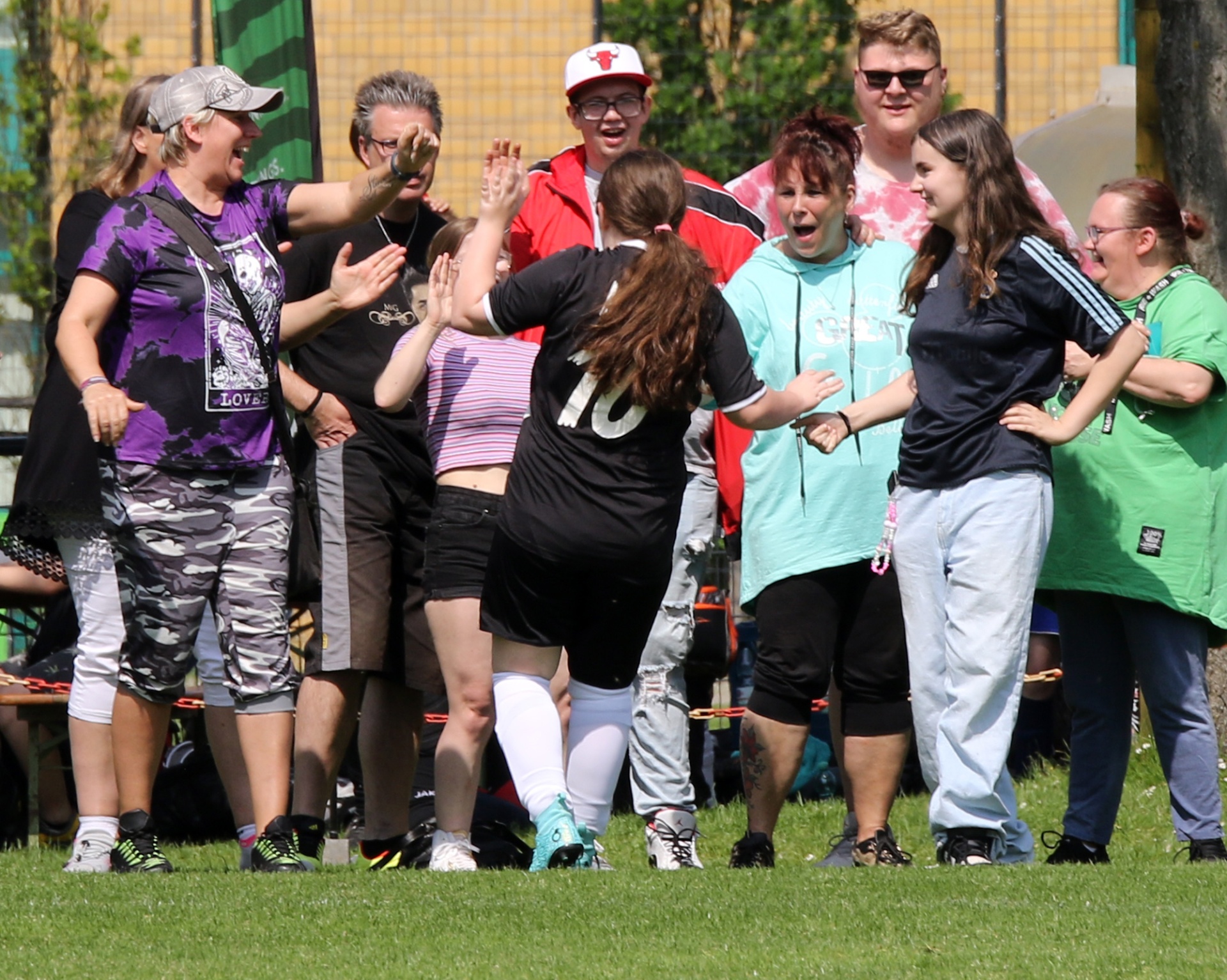 [{"clang_id":1,"value":"Gruppe Menschen auf einer Wiese, jubeln und geben sich High-Five. Verschiedene Freizeitkleidung, sonniger Tag. Im Hintergrund Gebäude und Bäume."}]