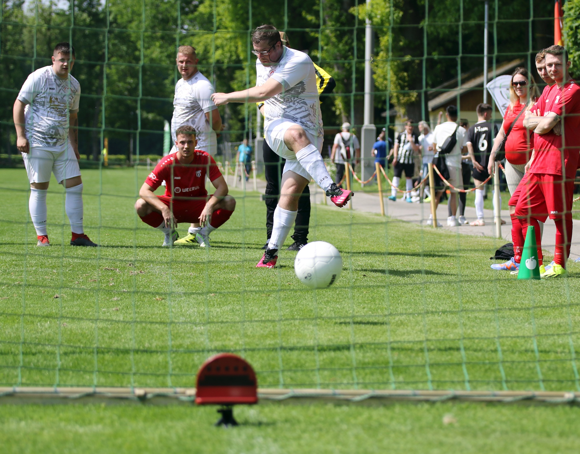 [{"clang_id":1,"value":"Fußballspieler schießt Ball auf Tor. Im Hintergrund weitere Spieler und Zuschauer am Spielfeldrand. Grüner Rasen und Zaun."}]