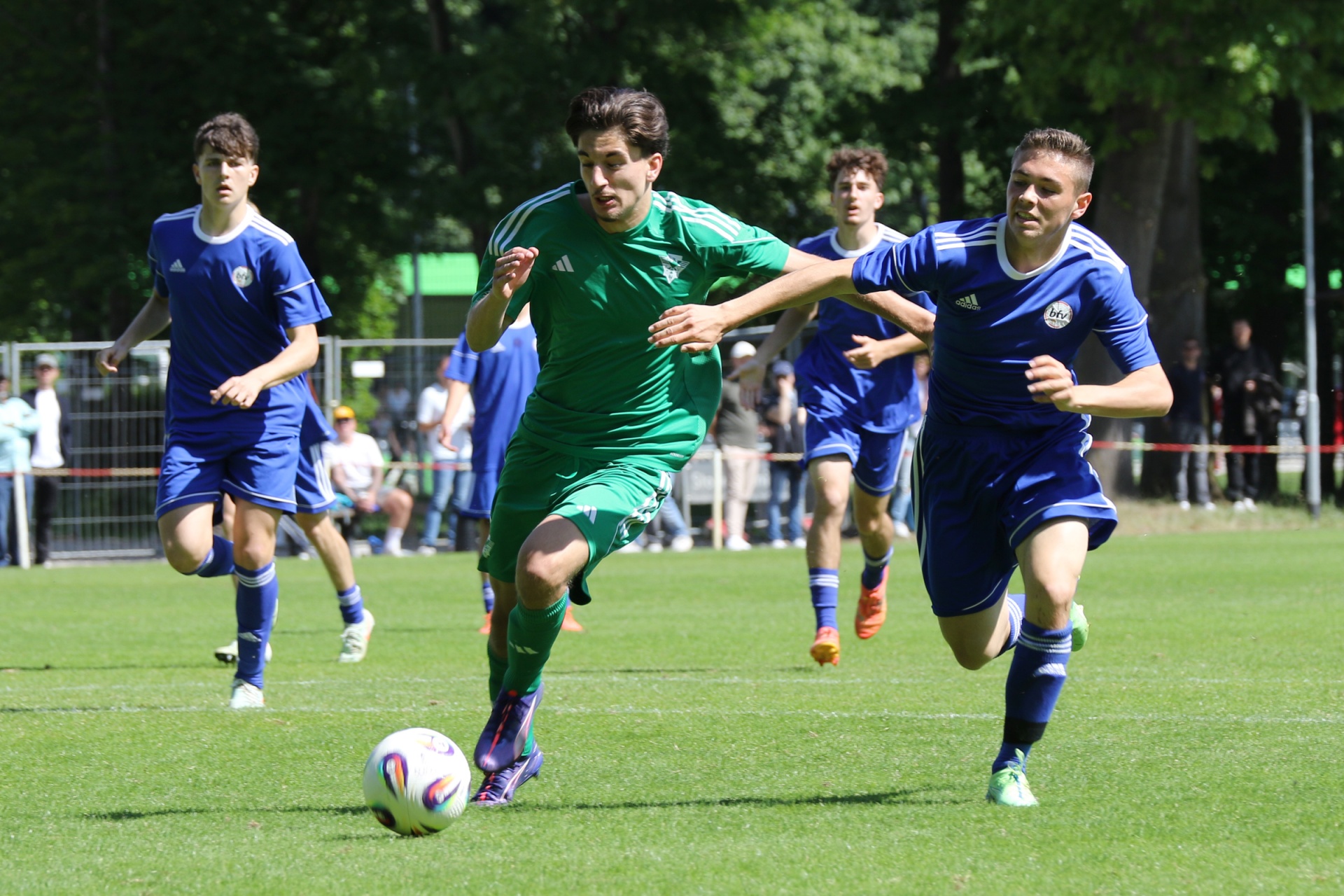 [{"clang_id":1,"value":"Fußballspiel: Zwei Spieler in grün und blau kämpfen um den Ball auf einem Rasenplatz. Im Hintergrund weitere Spieler und Zuschauer."}]