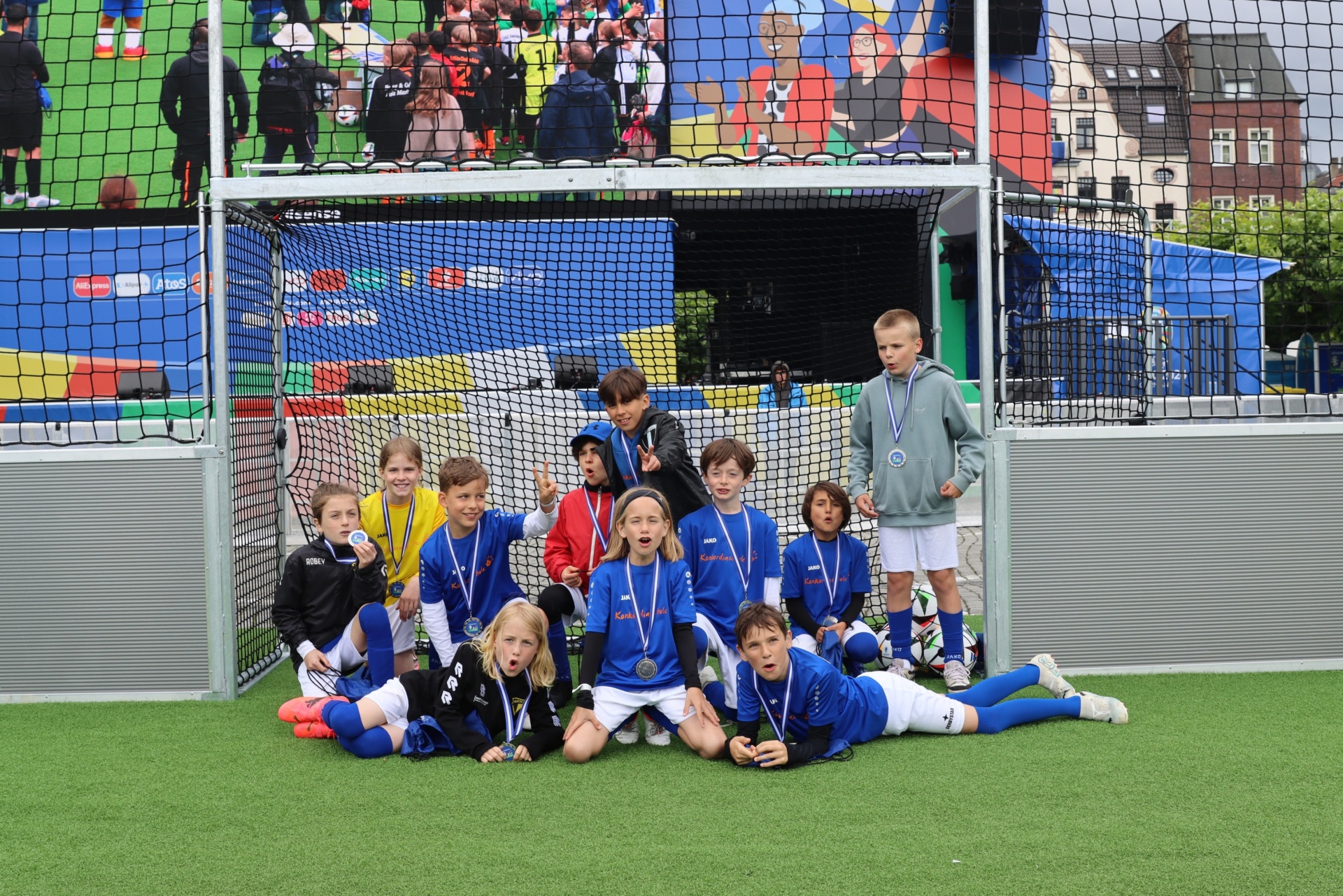 [{"clang_id":1,"value":"Fußballmannschaft mit Medaillen vor einem Fußballtor auf einem grünen Rasen. Im Hintergrund ein Stadion mit Zuschauern und Gebäuden."}]
