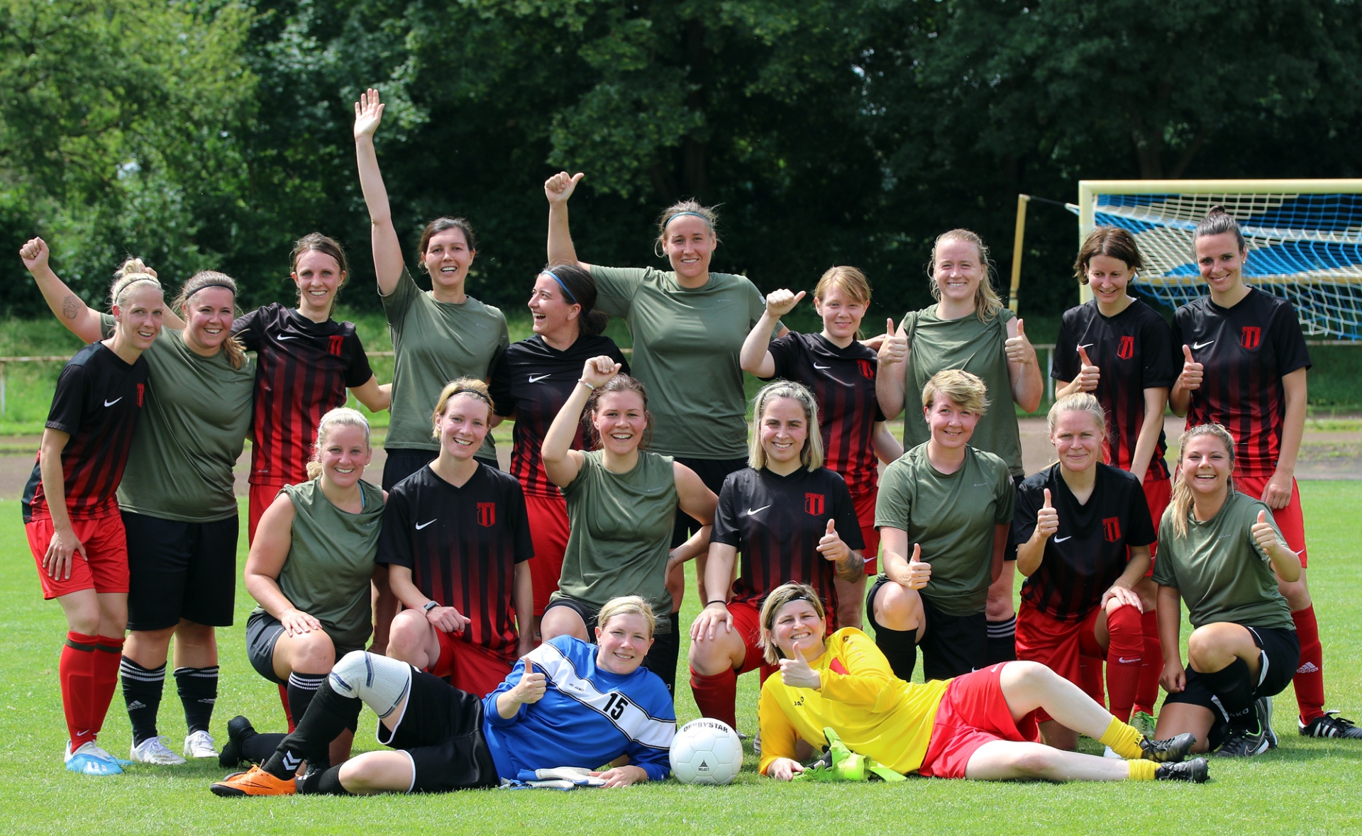 [{"clang_id":1,"value":"Frauenfußballteam in grünen und roten Trikots, einige sitzen oder liegen auf dem Rasen, lächeln und zeigen Daumen hoch. Im Hintergrund ein Fußballtor und Bäume."}]