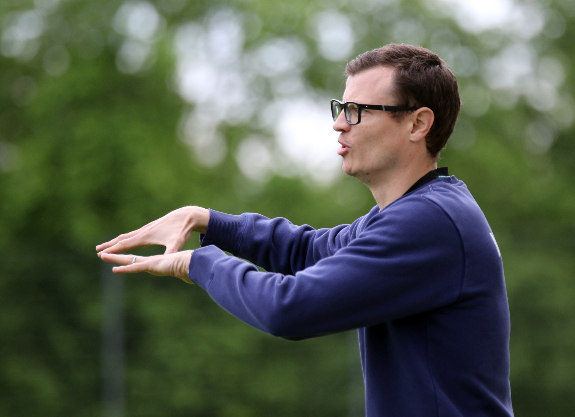 [{"clang_id":1,"value":"Mann mit Brille und dunklem Pullover gestikuliert im Freien, grüne Bäume im Hintergrund."}]