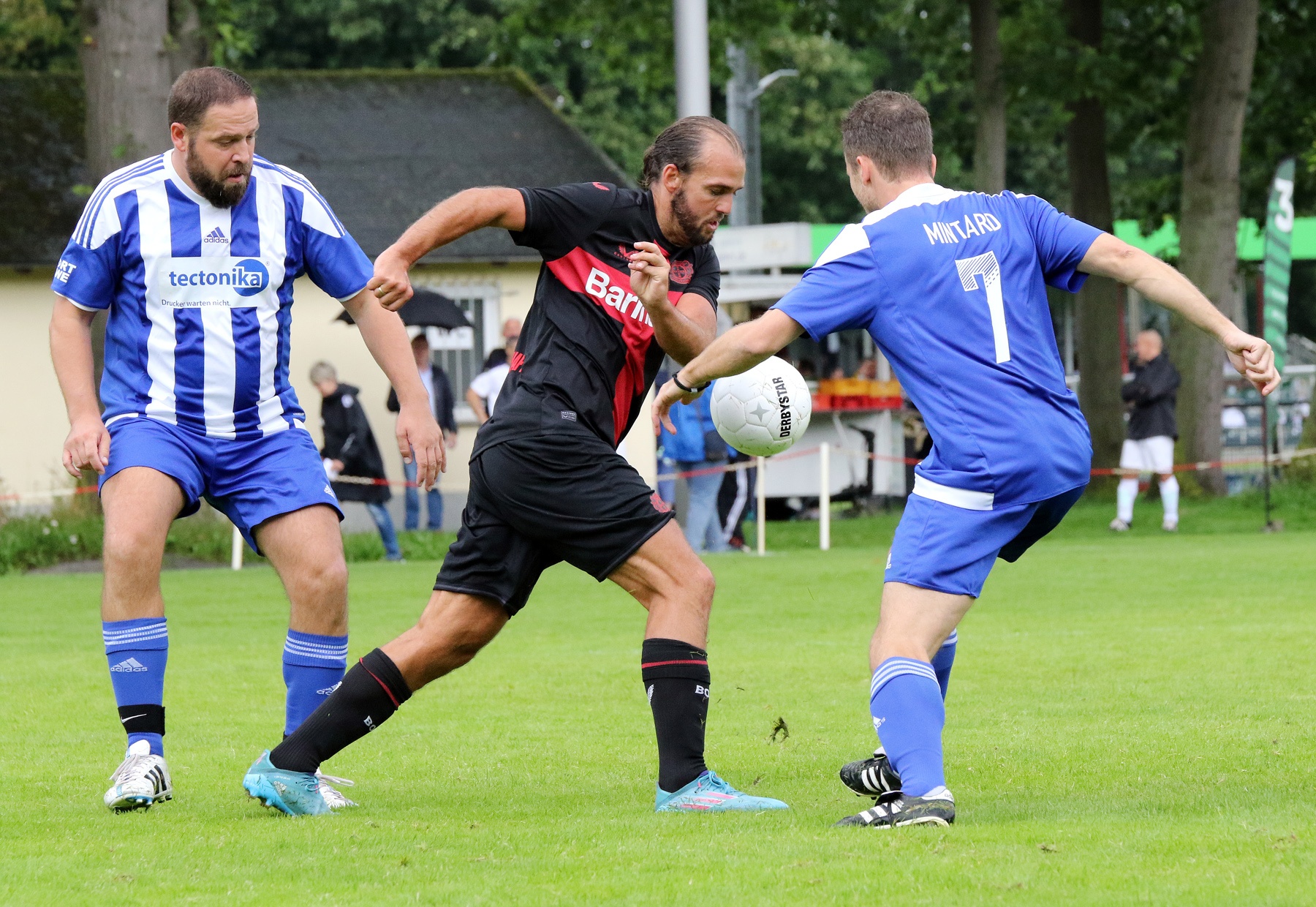 [{"clang_id":1,"value":"Drei Fußballspieler auf Grün. Links: Spieler in blau-weißem Trikot, Mitte: Spieler in rot-schwarzem Trikot mit Ball, rechts: Spieler in blauem Trikot. Hintergrund: Zuschauer und Bäume."}]