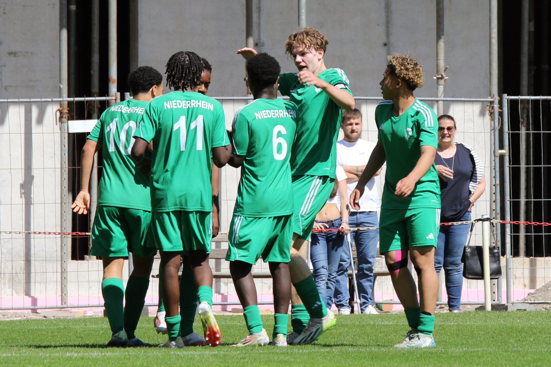 [{"clang_id":1,"value":"Fußballspieler in grünen Trikots mit Aufschrift \"NIEDERRHEIN\" feiern ein Tor. Im Hintergrund Zuschauer und eine Metallkonstruktion."}]
