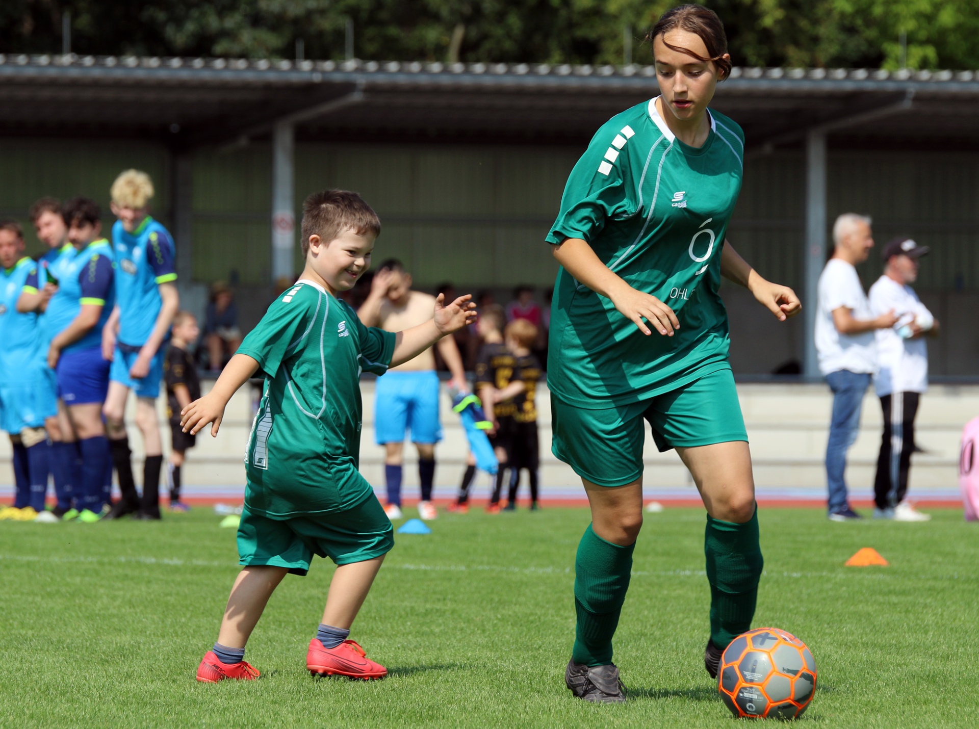 [{"clang_id":1,"value":"Fußballfeld: Frau in grün dribbelt Ball, Junge in grün rennt ihr zu. Im Hintergrund weitere Spieler und Zuschauer."}]