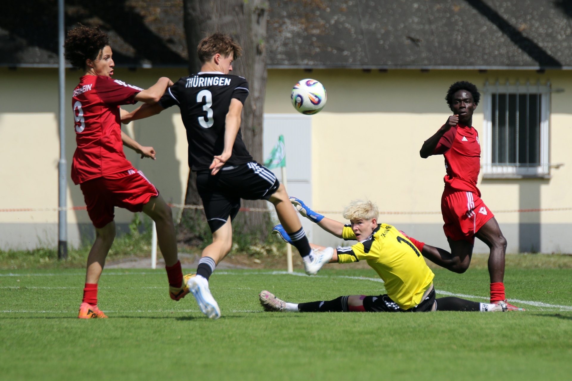 [{"clang_id":1,"value":"Fußballspiel: Spieler in Schwarz schießt auf Tor, Torwart in Gelb hält, zwei weitere Spieler in Rot im Hintergrund. Rasenplatz, Gebäude im Hintergrund."}]