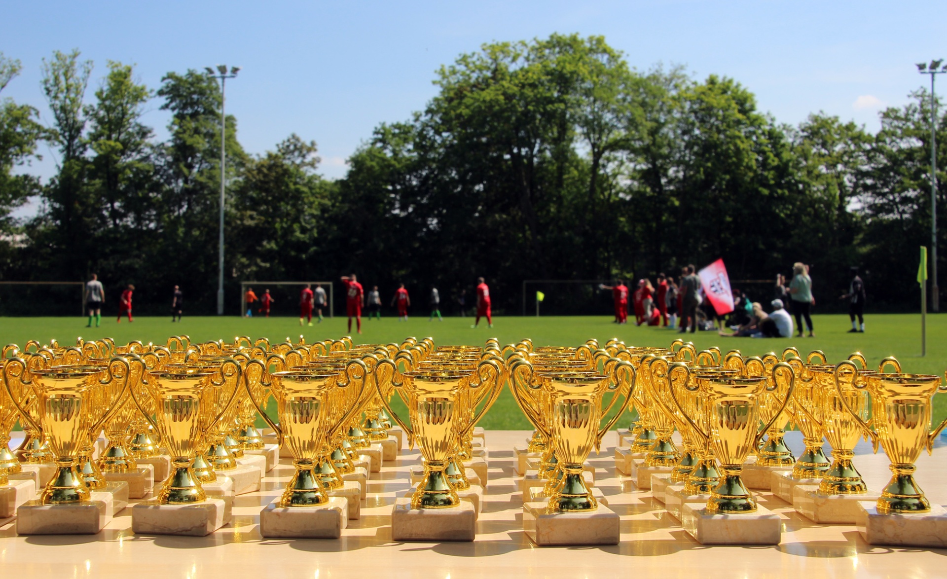 [{"clang_id":1,"value":"Goldene Pokale auf einer weißen Fläche im Vordergrund, dahinter Fußballfeld mit spielenden Menschen und Bäumen unter blauem Himmel."}]