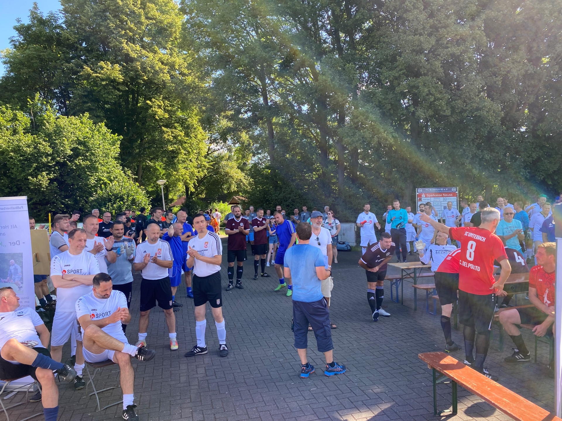 [{"clang_id":1,"value":"Fußballspieler in Sportkleidung stehen und sitzen auf einem Platz vor einem Gebäude mit Bäumen im Hintergrund. Einige applaudieren."}]