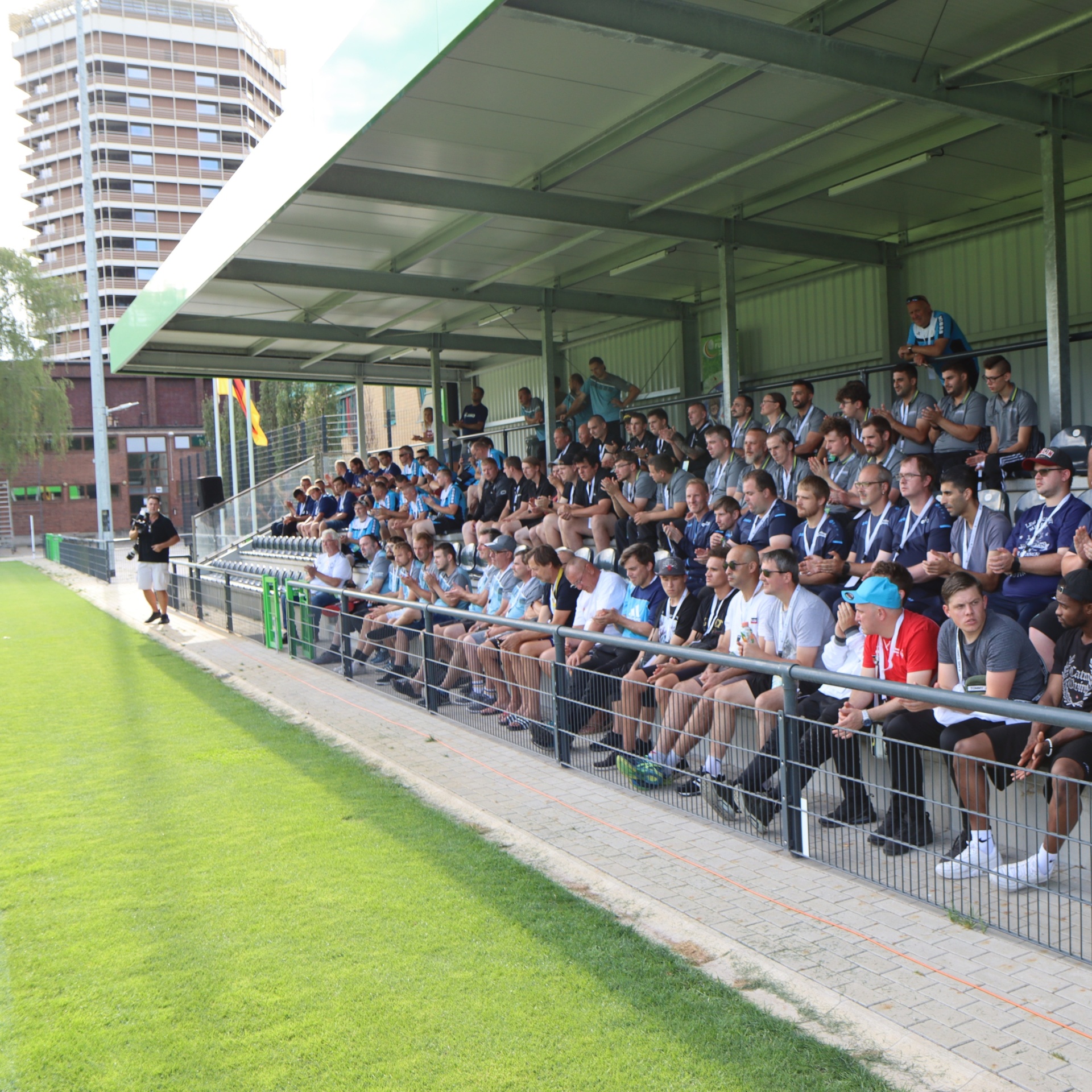[{"clang_id":1,"value":"Zuschauerreihen an einem Sportplatz, gefüllt mit Männern in Freizeitkleidung. Im Hintergrund ein Stadion und ein Hochhaus."}]