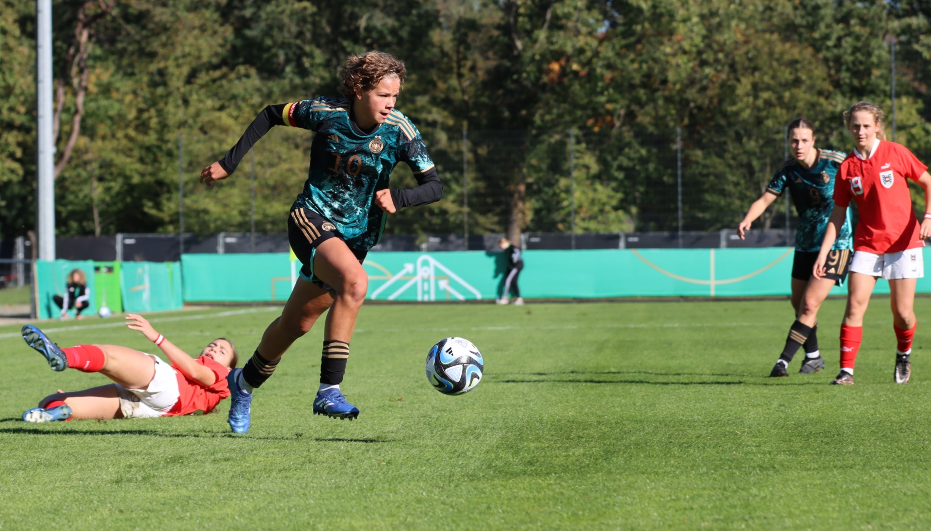 [{"clang_id":1,"value":"Fußballspiel: Spielerin in blau-grünem Trikot dribbelt den Ball, während eine Gegenspielerin in rot zu Boden geht. Im Hintergrund weitere Spielerinnen und grüne Bäume."}]