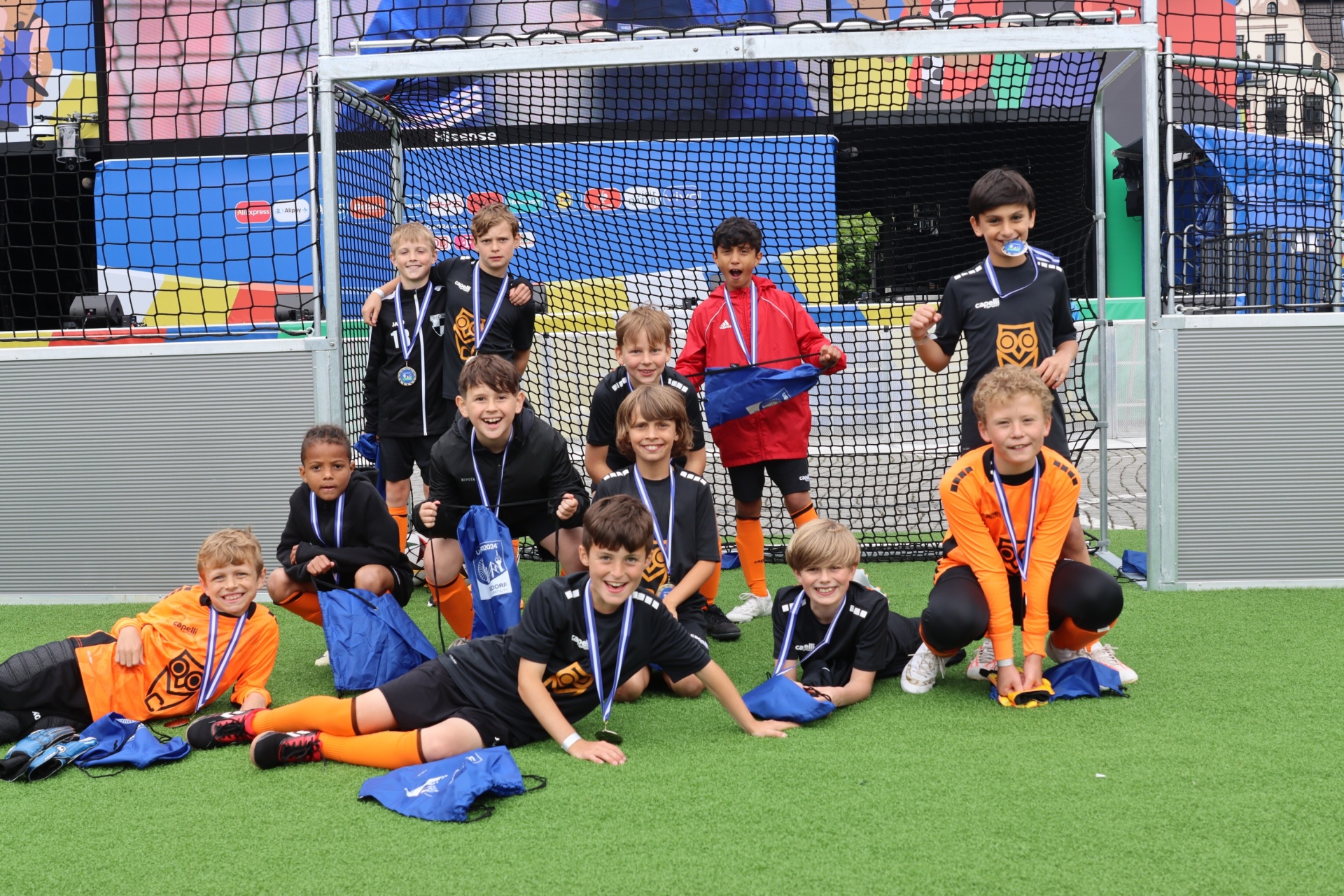 [{"clang_id":1,"value":"Fußballmannschaft mit Kindern posiert vor einem Fußballtor auf grünem Kunstrasen. Einige tragen Medaillen und halten Sporttaschen."}]
