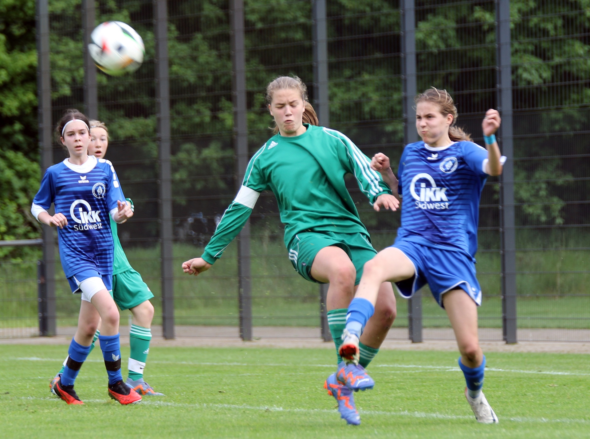 [{"clang_id":1,"value":"Drei Fußballspielerinnen auf grünem Rasen. Eine in Grün spielt den Ball, zwei in Blau stehen daneben. Im Hintergrund ein Zaun und Bäume."}]