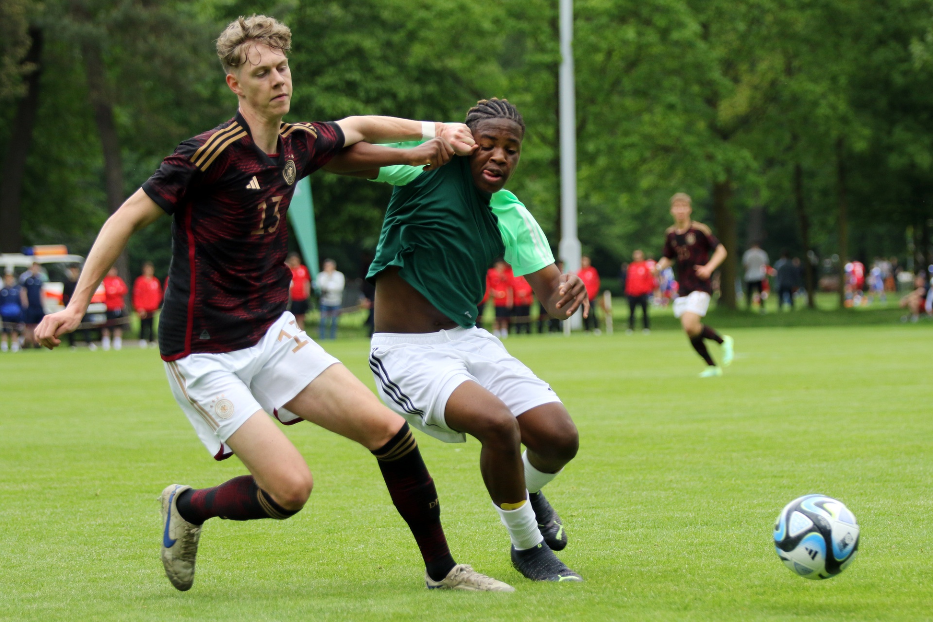 [{"clang_id":1,"value":"Zwei Fussballspieler im Wettkampf. Links ein Spieler mit blonden Haaren in dunkelrotem Trikot, rechts ein Spieler mit dunkler Haut in grünem Trikot. Ein Fussball liegt auf dem Rasen."}]