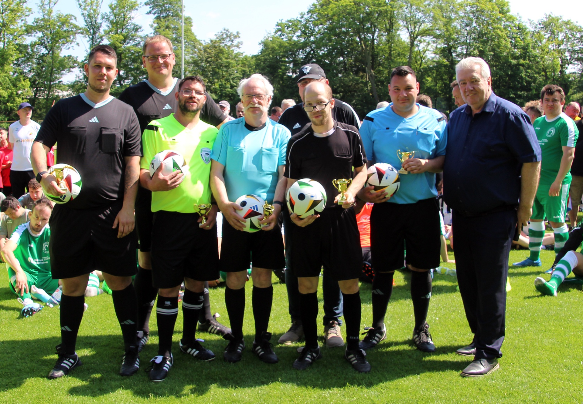 [{"clang_id":1,"value":"Gruppe von acht Männern auf einem Fußballfeld, einige in Schiedsrichteruniform, mit Pokalen und Fußbällen. Im Hintergrund spielende Fußballspieler."}]