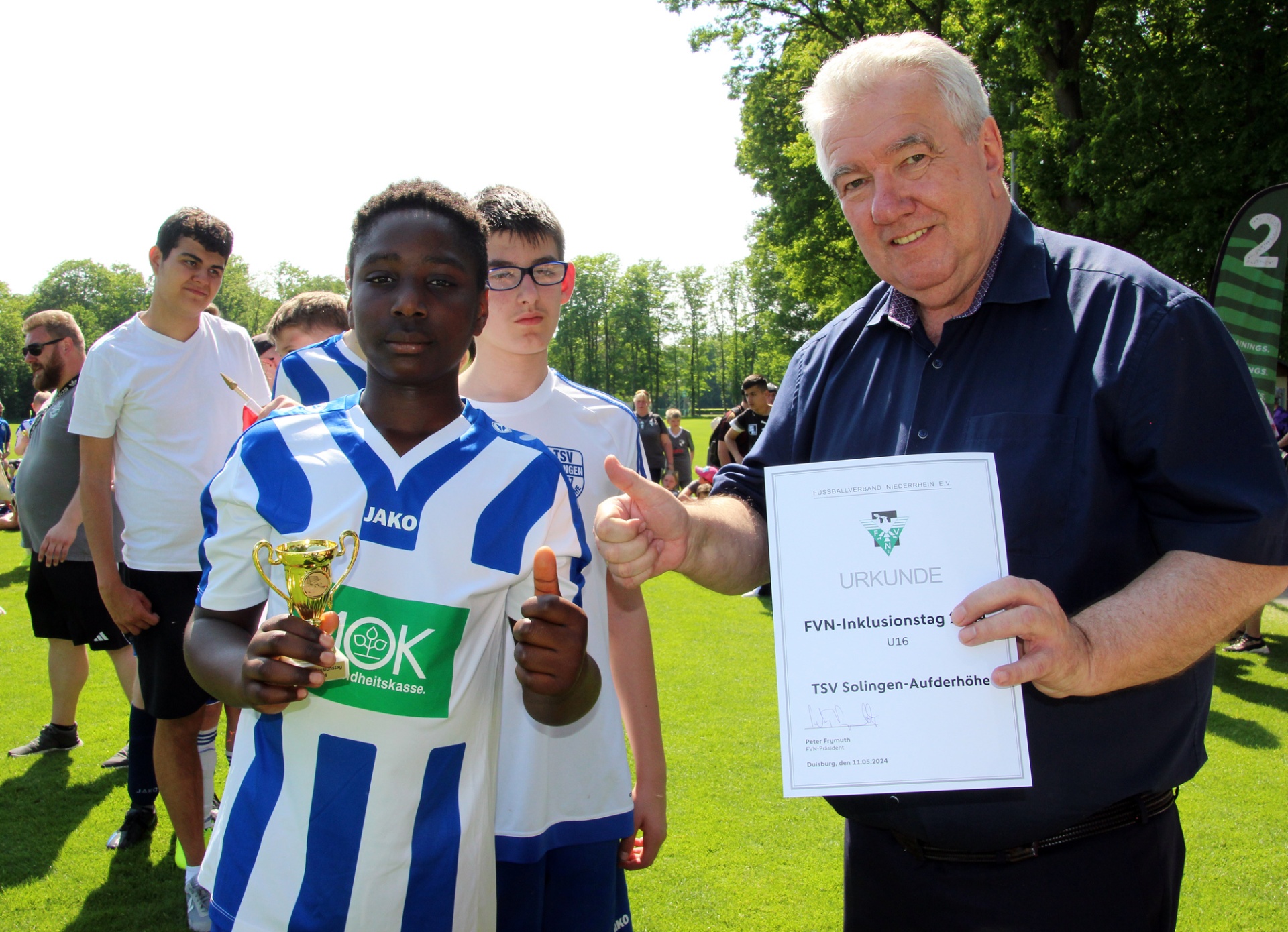[{"clang_id":1,"value":"Fußballturnier: Drei Jugendliche mit Pokal und Urkunde, überreicht von einem älteren Mann. Rasen und Bäume im Hintergrund."}]