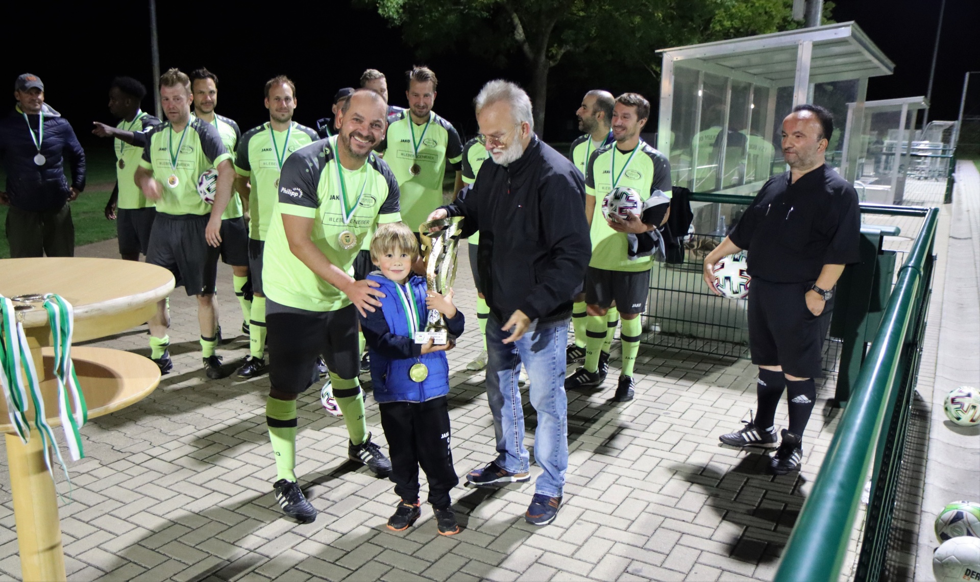[{"clang_id":1,"value":"Fußballteam in grünen Trikots mit Medaillen und Pokal posiert vor einem Fußballfeld bei Nacht. Ein Mann überreicht einem kleinen Jungen eine Medaille."}]