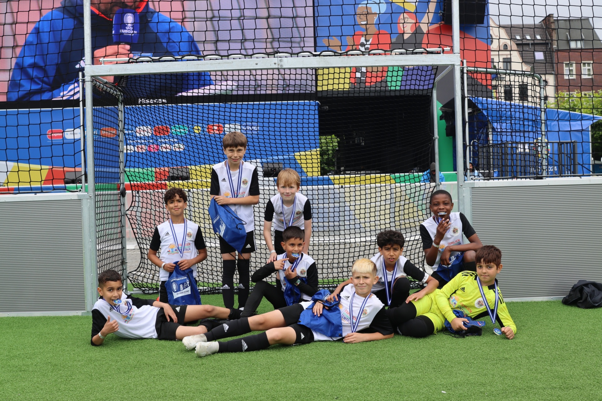 [{"clang_id":1,"value":"Fußballmannschaft mit Kindern, alle tragen Medaillen und Sportkleidung, sitzt vor einem Fußballtor auf einem Kunstrasenplatz. Im Hintergrund Häuser."}]