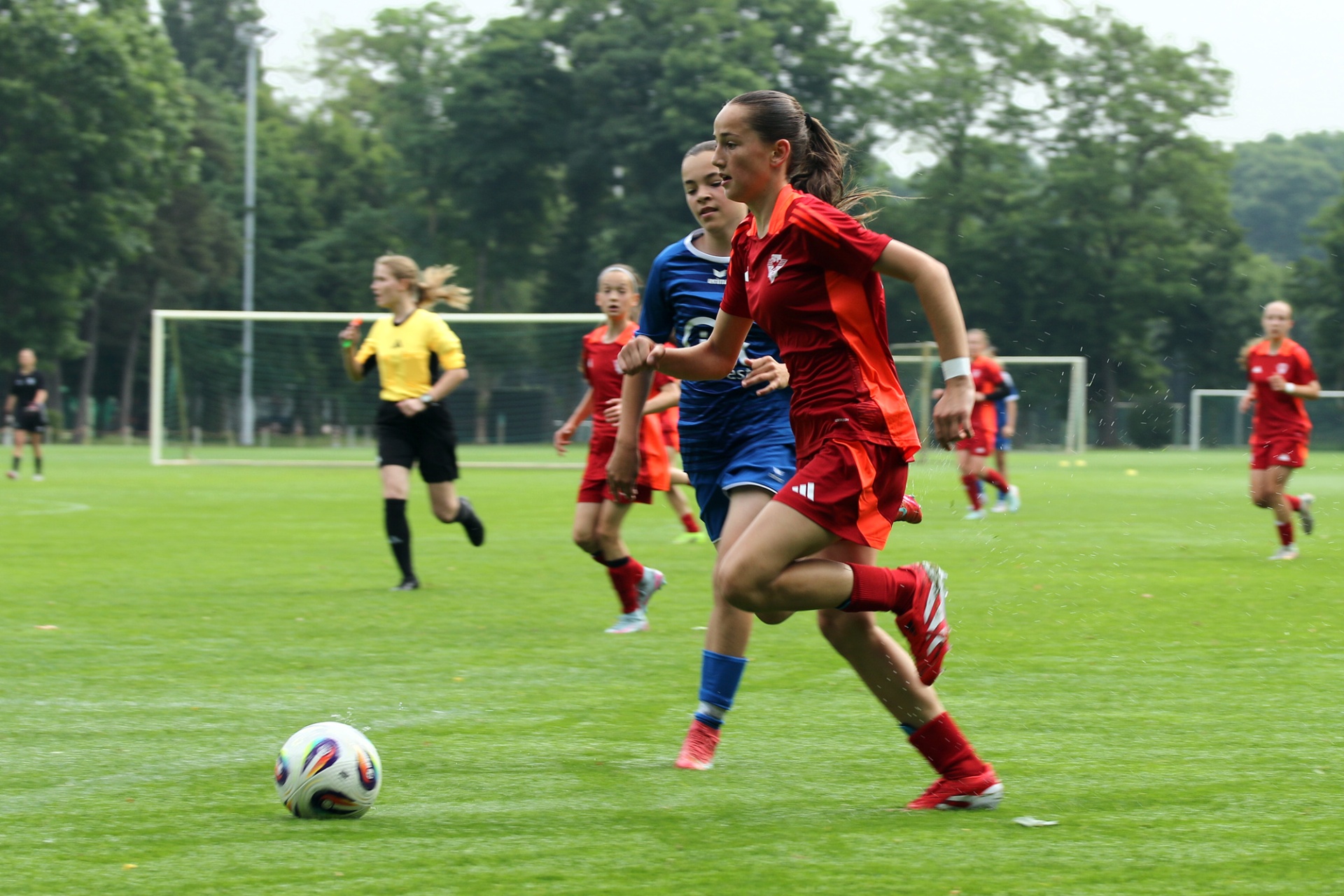 [{"clang_id":1,"value":"Fußballspiel: Eine Spielerin in rotem Trikot läuft mit Ball auf dem grünen Rasen. Im Hintergrund weitere Spielerinnen und das Spielfeld."}]