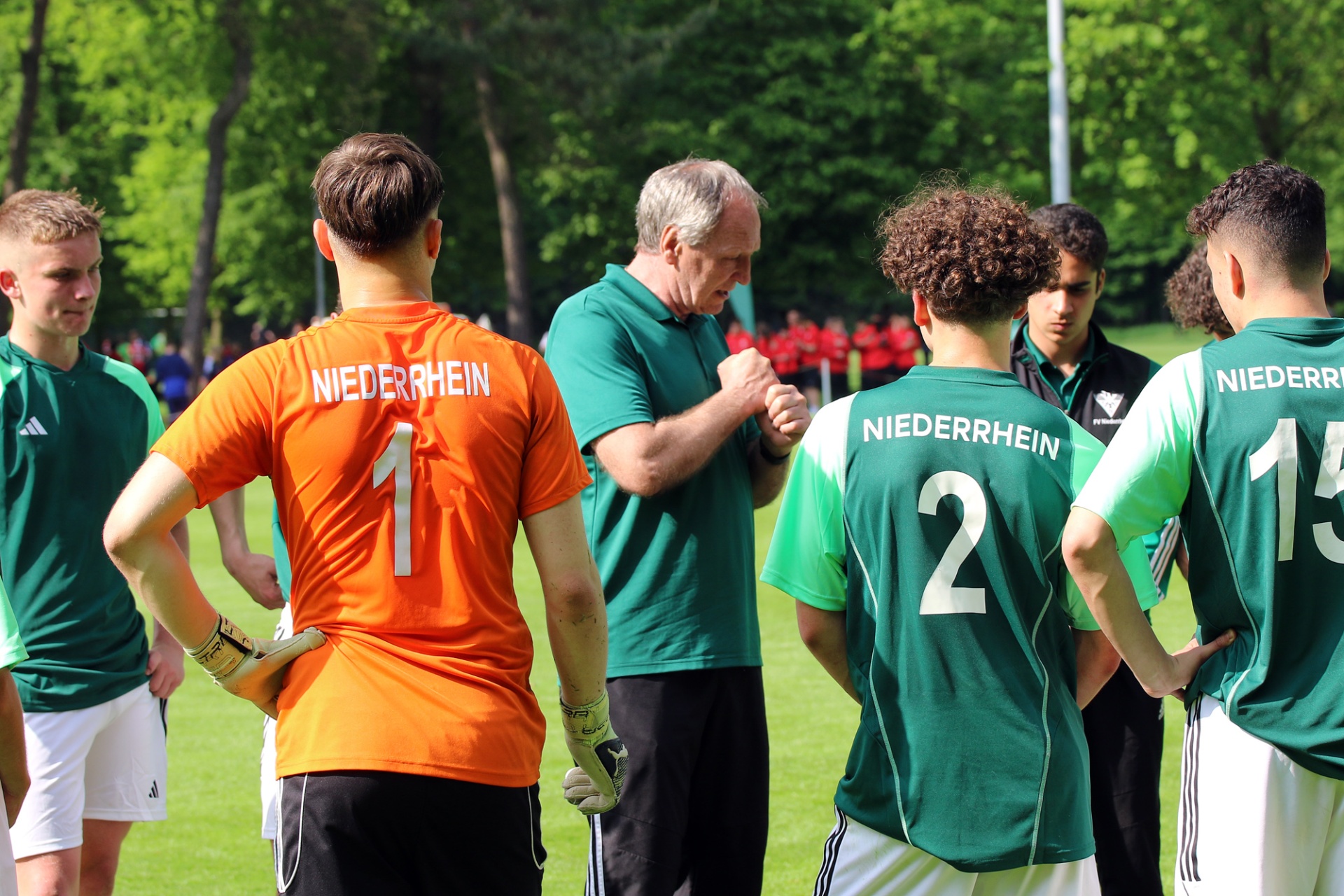 [{"clang_id":1,"value":"Fußballspieler mit Trikots der Nummern 1, 2 und 13 stehen im Kreis um einen Trainer. Hintergrund: Bäume und Grünflächen."}]