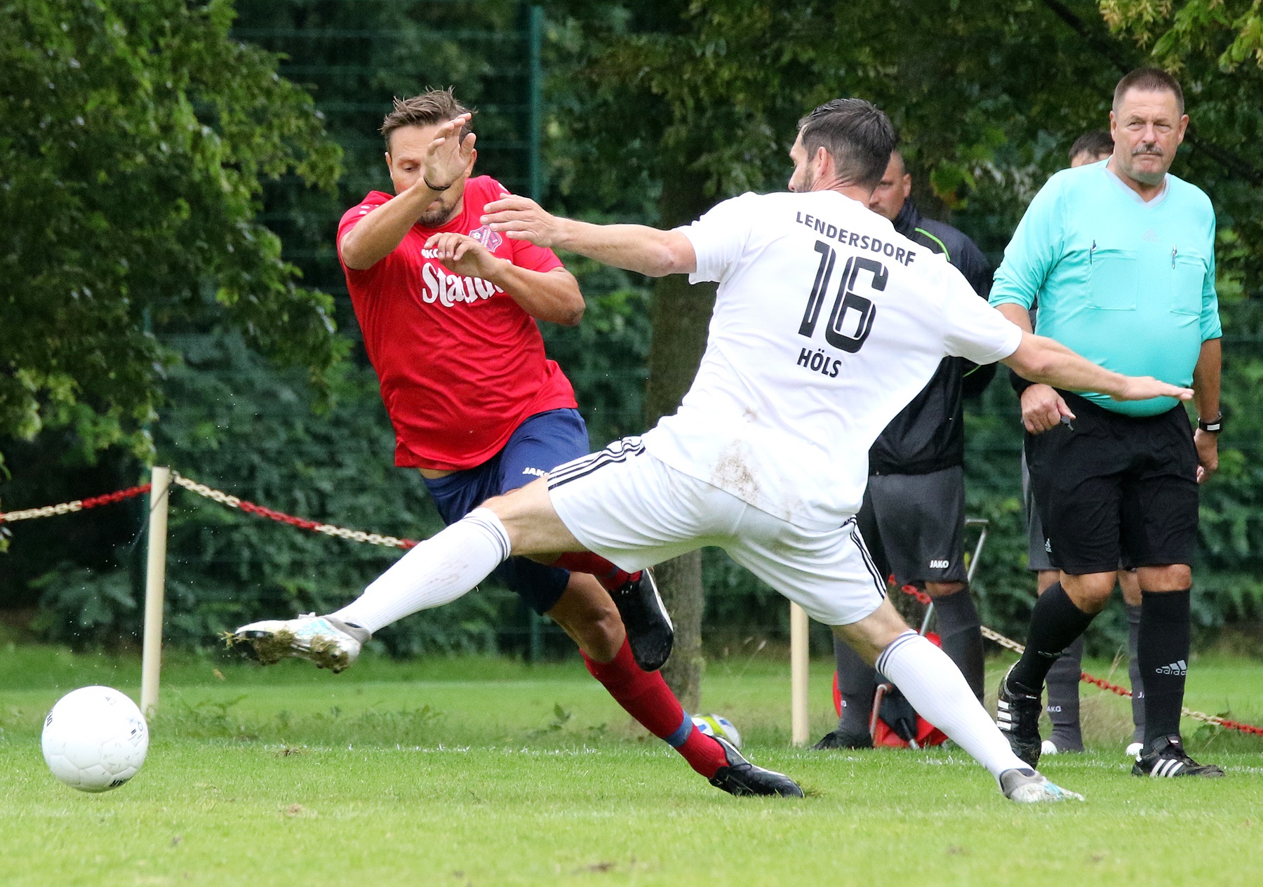 [{"clang_id":1,"value":"Zwei Fußballspieler kämpfen um den Ball. Links ein Spieler in Rot, rechts in Weiß mit der Aufschrift \"LENDERSDORE 16\". Im Hintergrund ein Schiedsrichter."}]