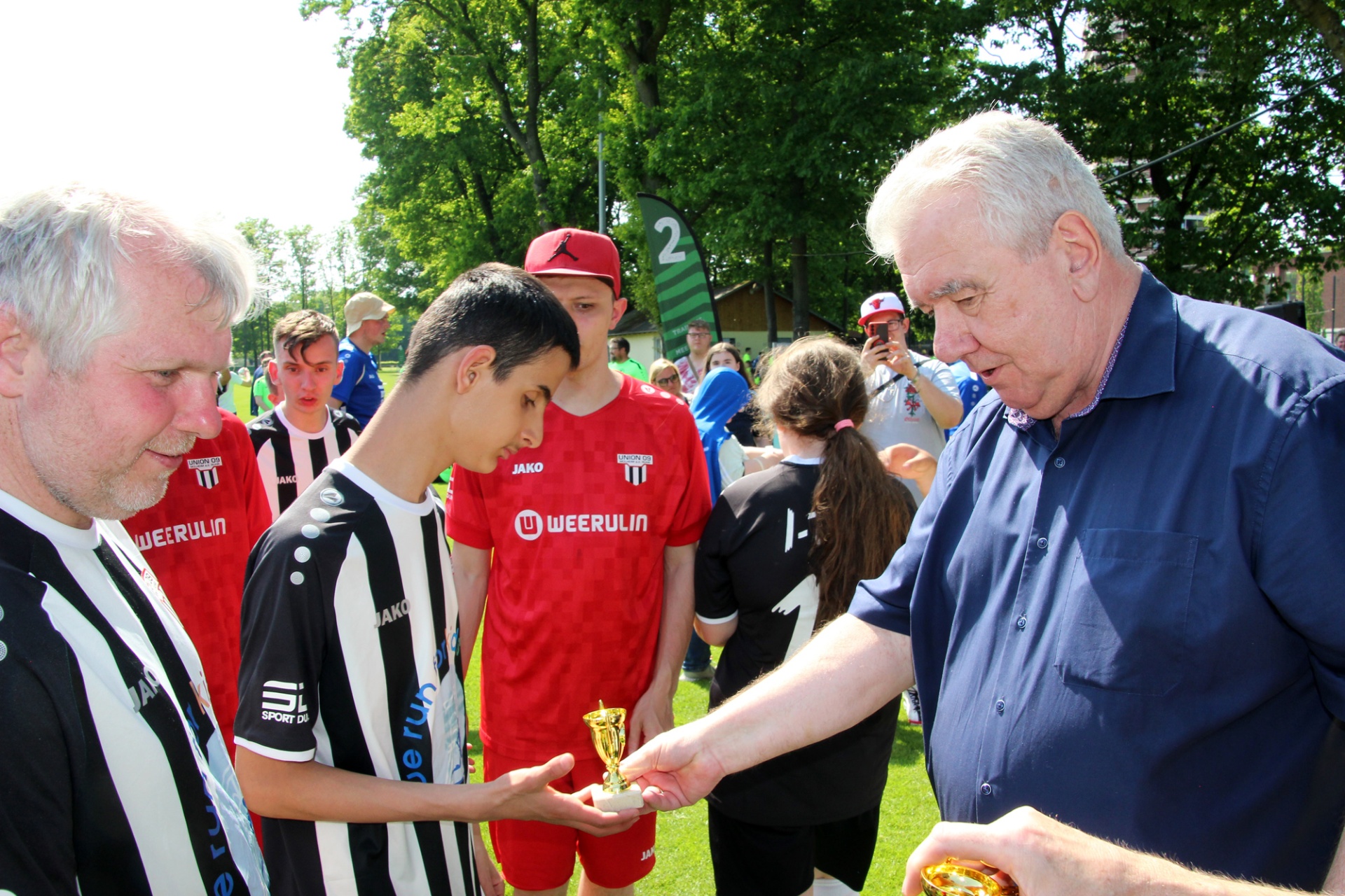 [{"clang_id":1,"value":"Zwei ältere Männer überreichen einem Jugendlichen in Fußballkleidung Pokale auf einem grünen Rasen. Im Hintergrund weitere Personen und Bäume."}]