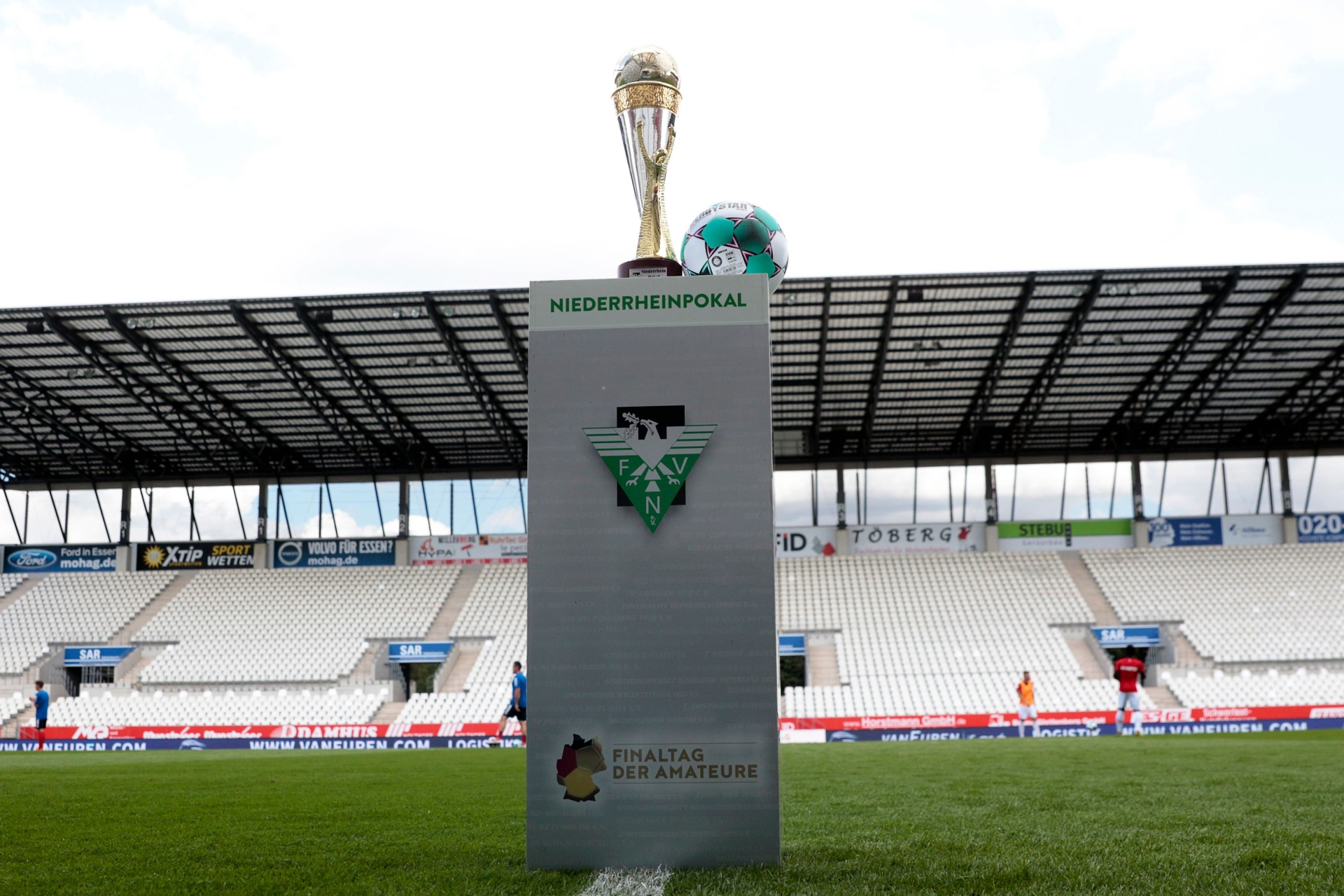 [{"clang_id":1,"value":"Pokal Niederrhein: Pokal auf Sockel vor Fußballstadion mit Zuschauerrängen und Werbebanden. Himmel ist bewölkt."}]