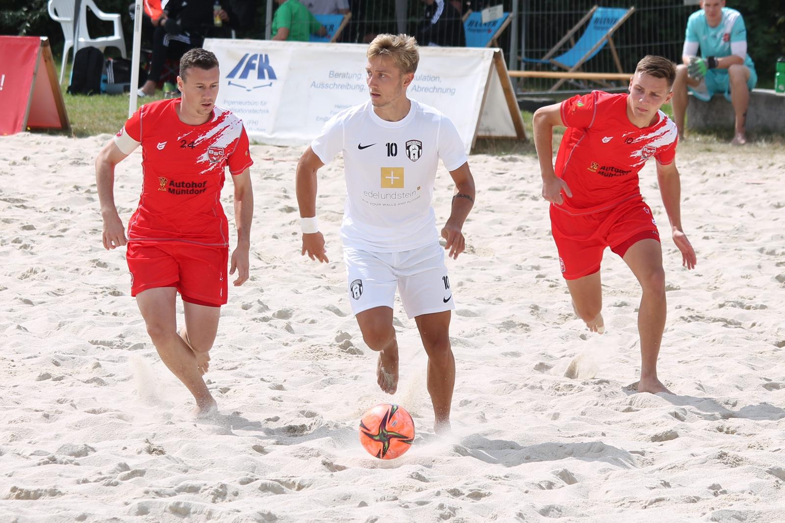 [{"clang_id":1,"value":"Drei Fussballspieler auf Sandstrand. Mittlerer Spieler in weissem Trikot mit Ball, links und rechts jeweils ein Spieler in roten Trikots. Im Hintergrund Zuschauer und Werbebanner."}]
