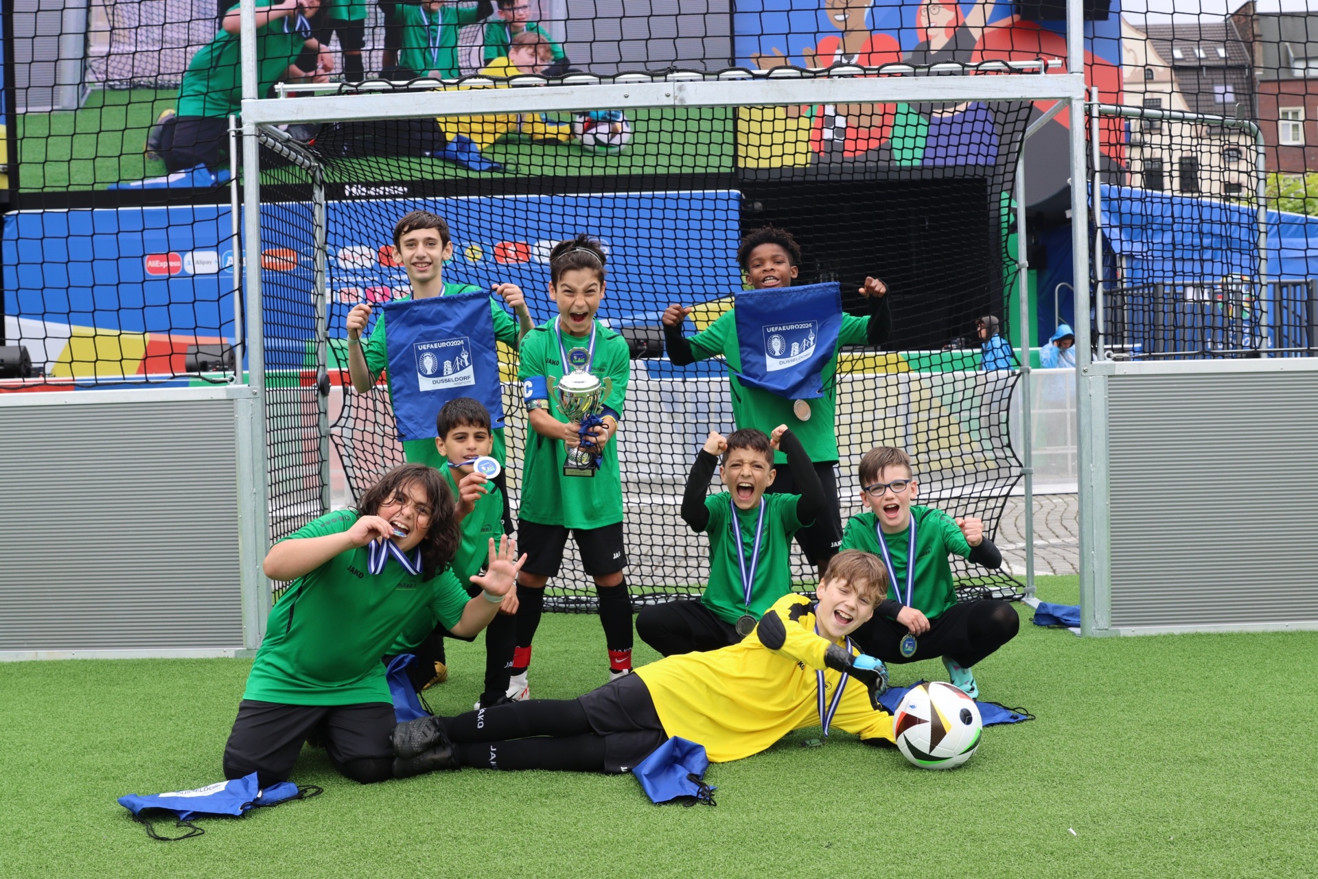 [{"clang_id":1,"value":"Fußballmannschaft mit Pokal und Medaillen posiert vor einem Fußballtor auf einem grünen Kunstrasenplatz. Sieben Jungen im Alter von ca. 10-14 Jahren."}]
