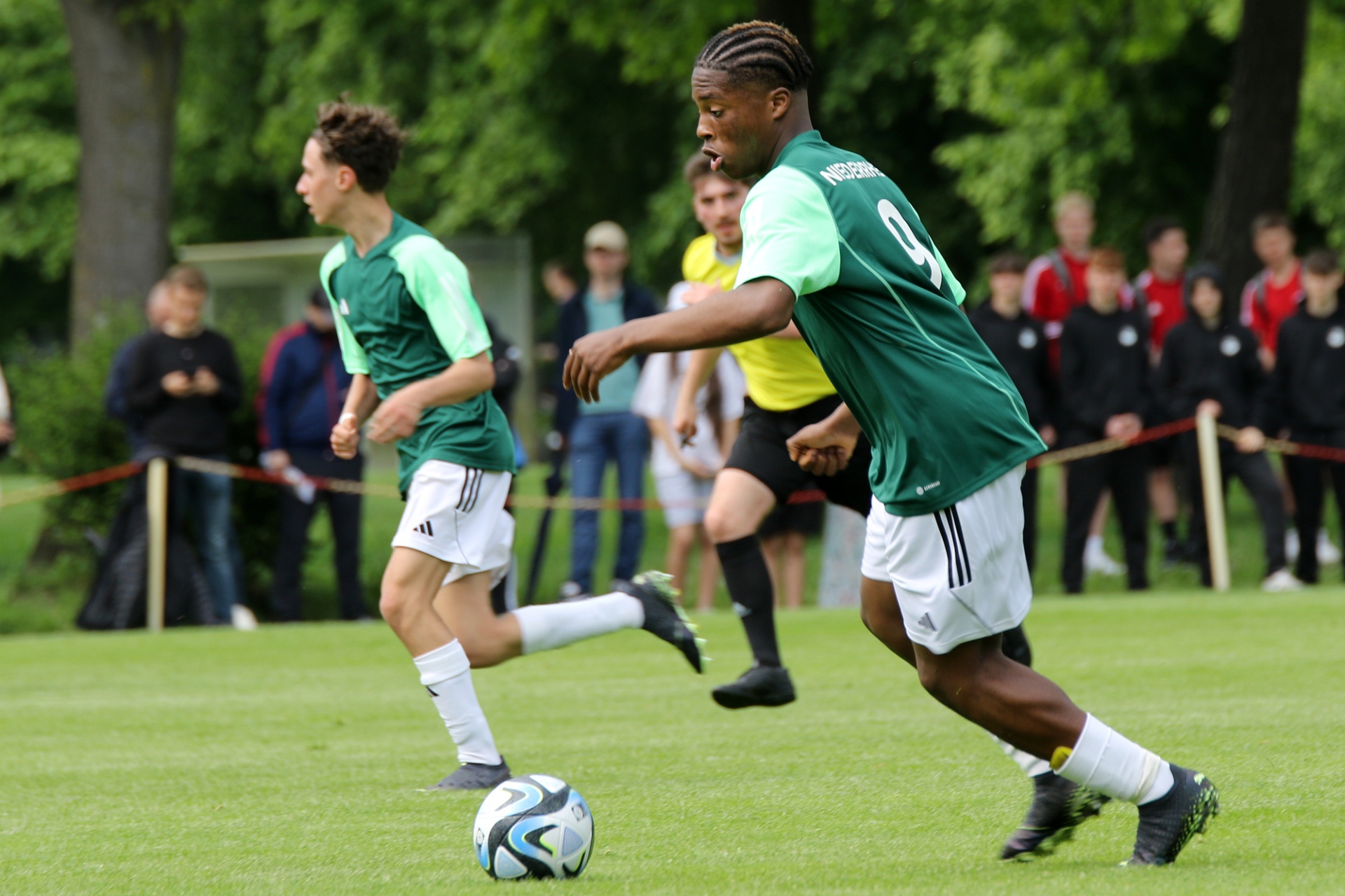 [{"clang_id":1,"value":"Zwei Fußballspieler in grünen Trikots und weißen Shorts auf Rasenfeld. Im Hintergrund Zuschauer und Bäume. Ein Spieler dribbelt den Ball."}]