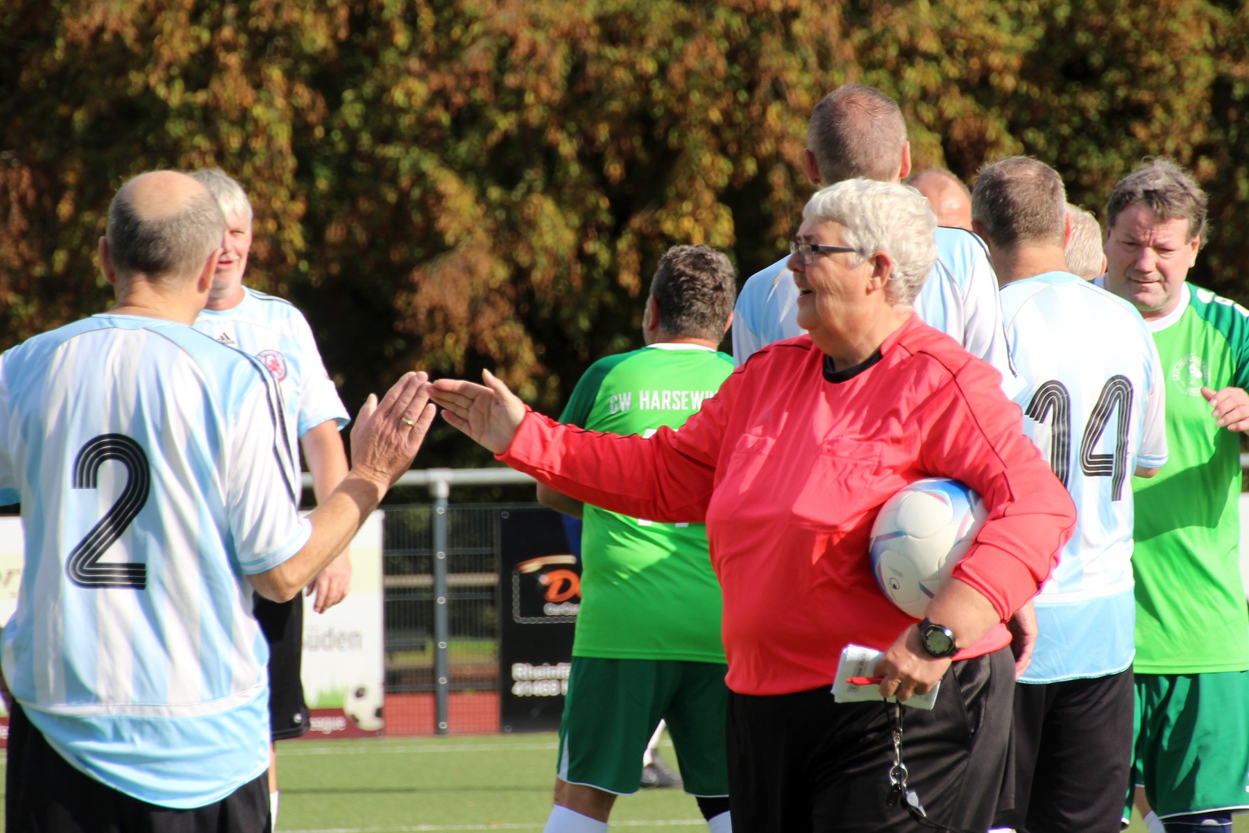 [{"clang_id":1,"value":"Fußballspieler in hellblauen und grünen Trikots stehen um einen Schiedsrichter in roter Jacke. Im Hintergrund Bäume und ein Fußballfeld."}]