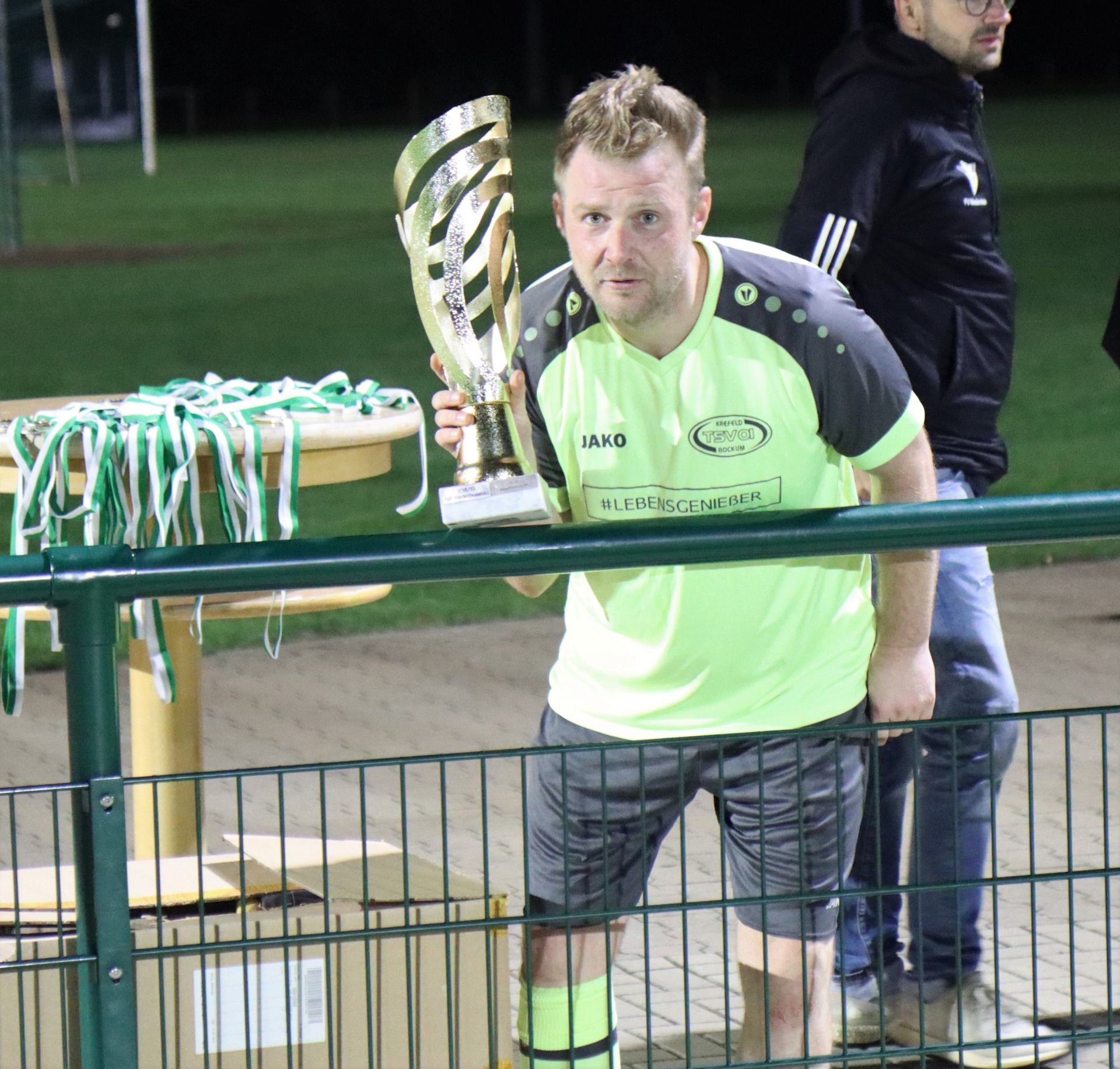 [{"clang_id":1,"value":"Mann mit Pokal. Er trägt ein grünes und gelbes Sporttrikot, dunkle Shorts und steht an einem Fußballfeld hinter einem Zaun. Im Hintergrund weitere Personen."}]