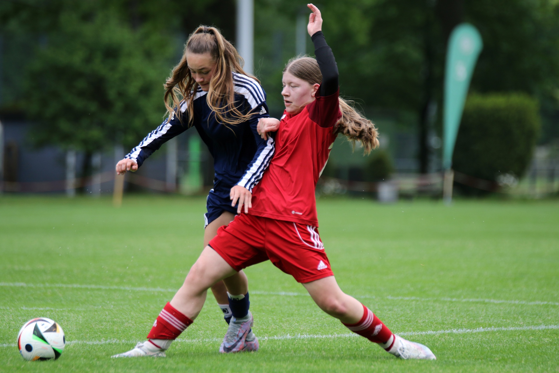 [{"clang_id":1,"value":"Zwei Fußballspielerinnen kämpfen um den Ball auf einem grünen Rasen. Eine trägt rot-weißes, die andere blau-weißes Trikot. Ein Fußball liegt im Vordergrund."}]