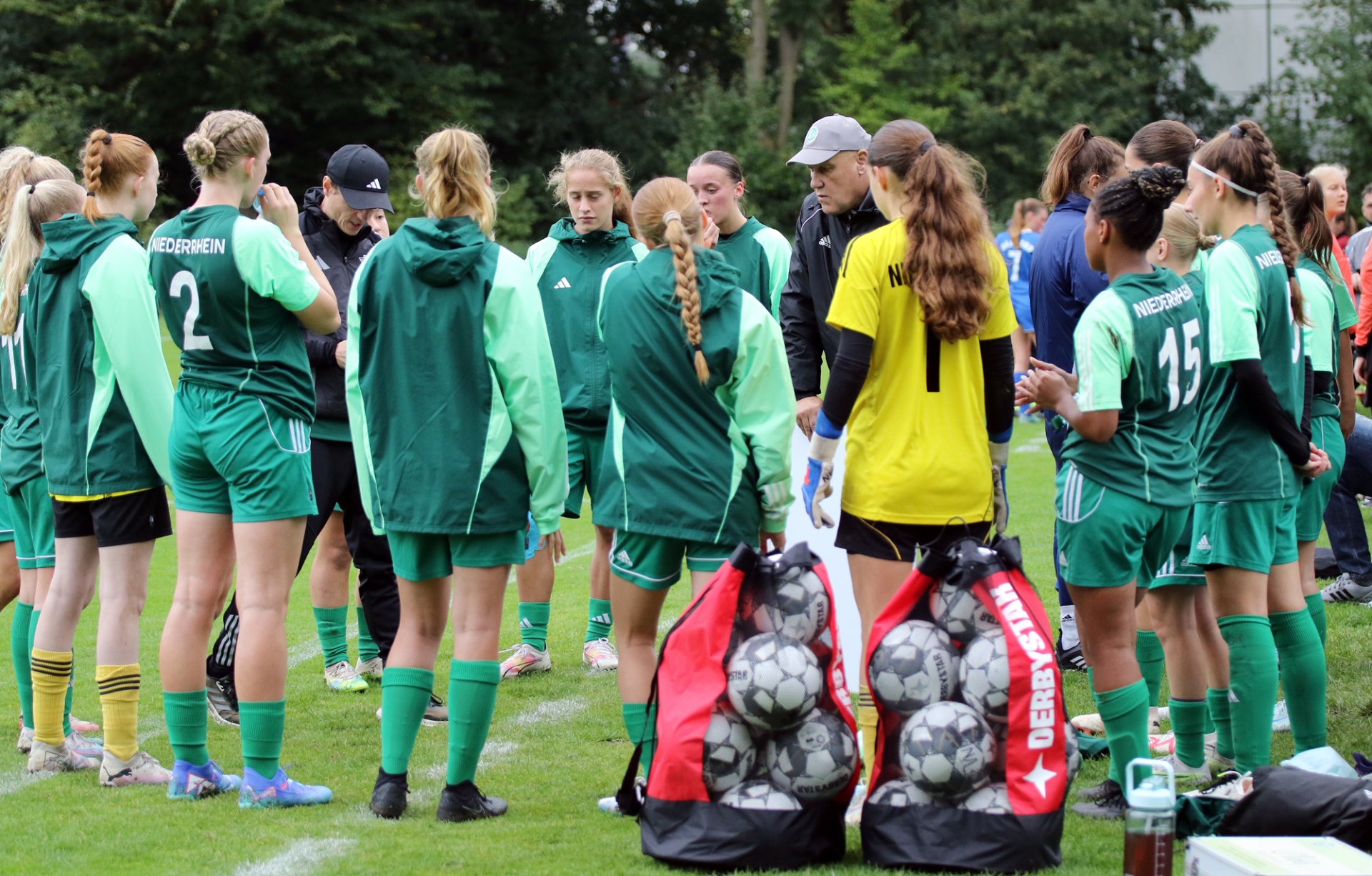 [{"clang_id":1,"value":"Fußballmannschaft in grünen Trikots steht auf Rasenfeld zusammen. Trainer gibt Anweisungen, Balltaschen in der Mitte."}]