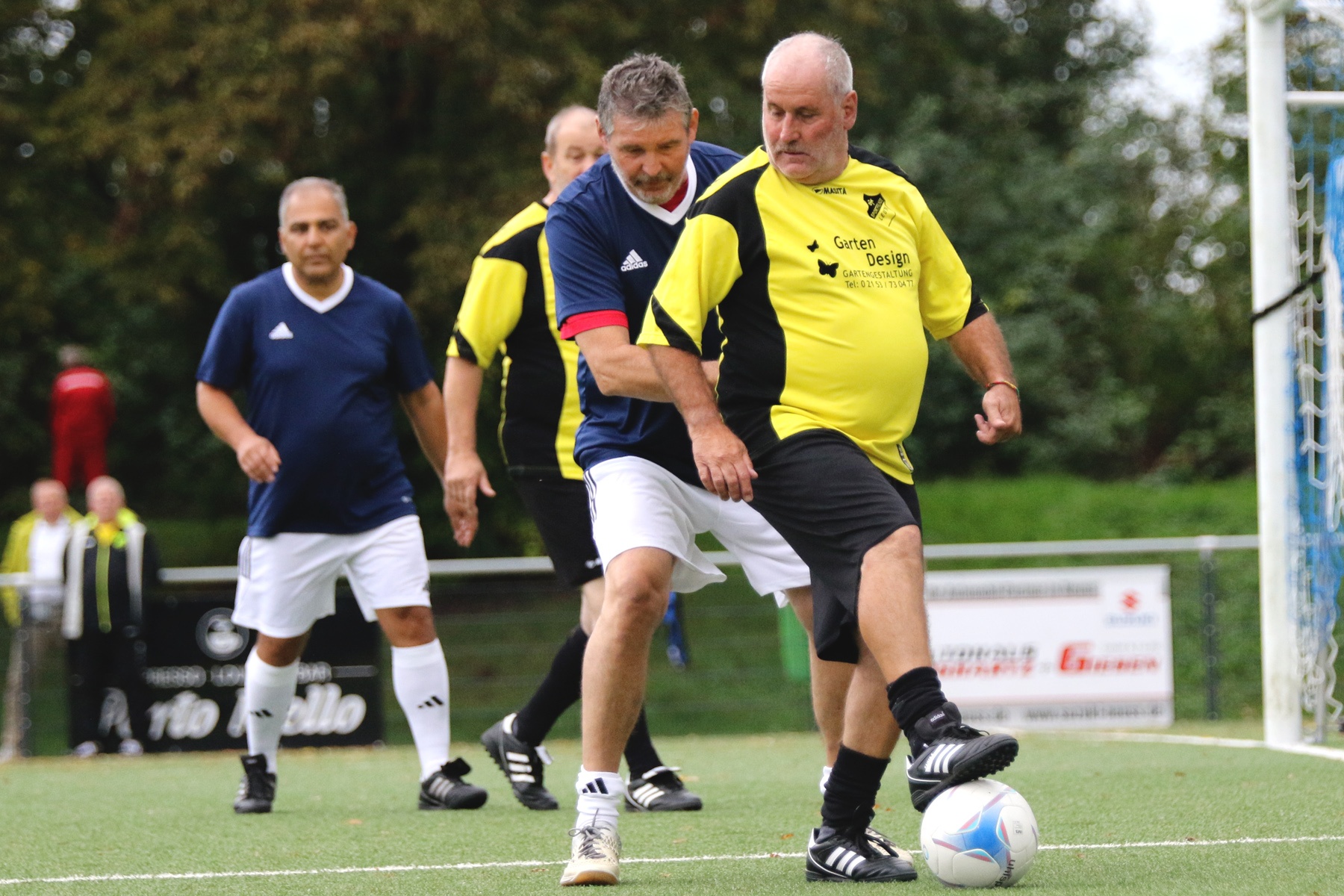 [{"clang_id":1,"value":"Drei Männer mittleren Alters spielen Fußball auf einem Rasenplatz. Ein Mann in Gelb dribbelt den Ball, ein anderer in Blau versucht ihn zu blocken. Im Hintergrund ein dritter Spieler und eine Bande."}]