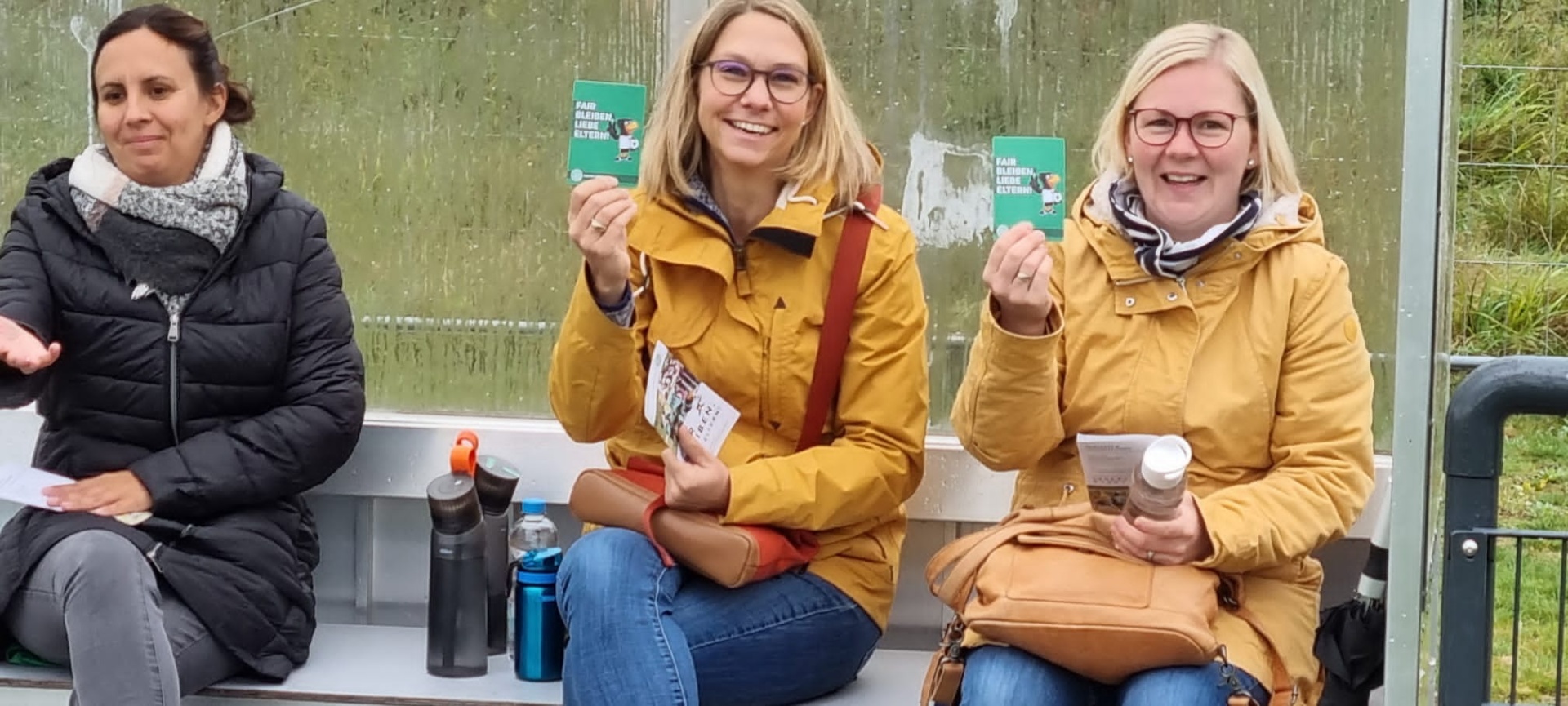 [{"clang_id":1,"value":"Drei lächelnde Frauen sitzen auf einer Bank vor einer grauen Wand. Sie halten jeweils eine grüne Karte in der Hand. Eine trägt eine dunkle Jacke, die anderen gelbe Mäntel."}]