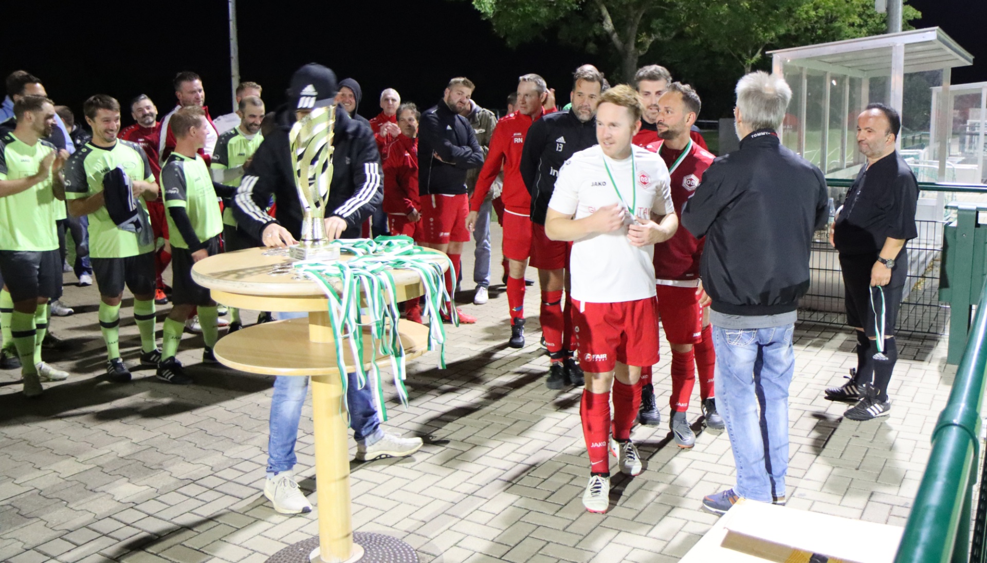 [{"clang_id":1,"value":"Fußballspieler in Vereinskleidung feiern um einen Tisch mit Pokal und Girlande. Einige stehen, andere sitzen auf einer Bank. Dunkler Hintergrund."}]