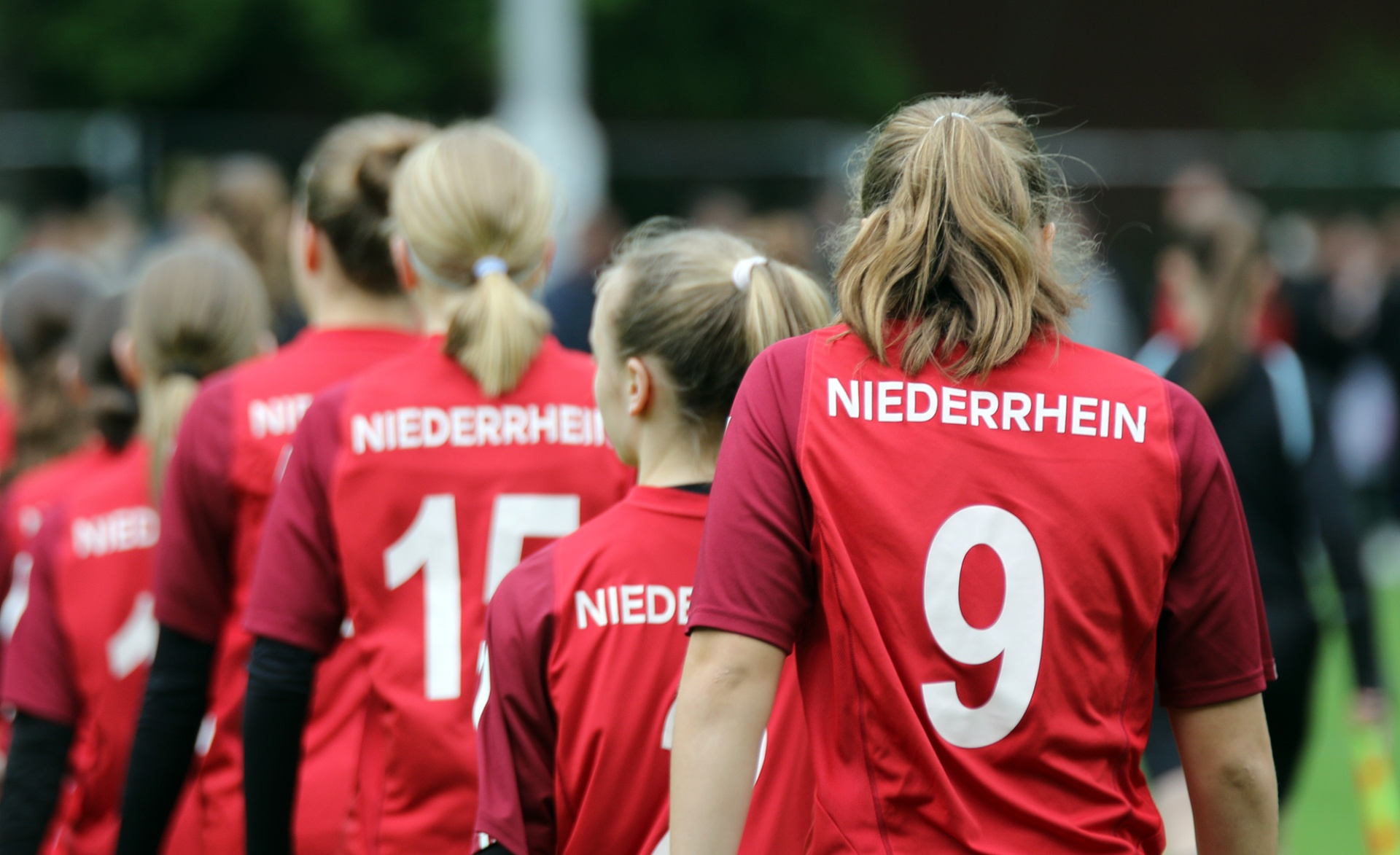 [{"clang_id":1,"value":"Fußballspielerinnen in roten Trikots mit Aufschrift \"NIEDERRHEIN\" und Nummern 9 und 17 stehen zusammen auf einem Fußballfeld."}]