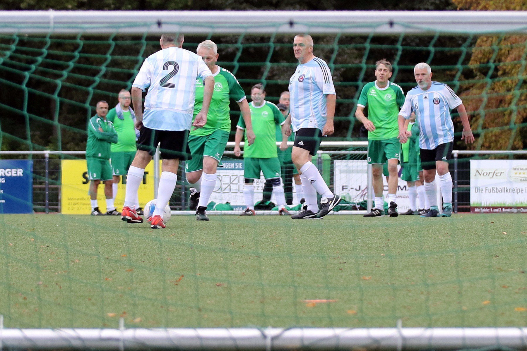 [{"clang_id":1,"value":"Fußballspiel: Zwei Teams stehen sich gegenüber, eines trägt grüne, das andere hellblaue Trikots. Im Hintergrund eine grüne Hecke und Werbebanner."}]