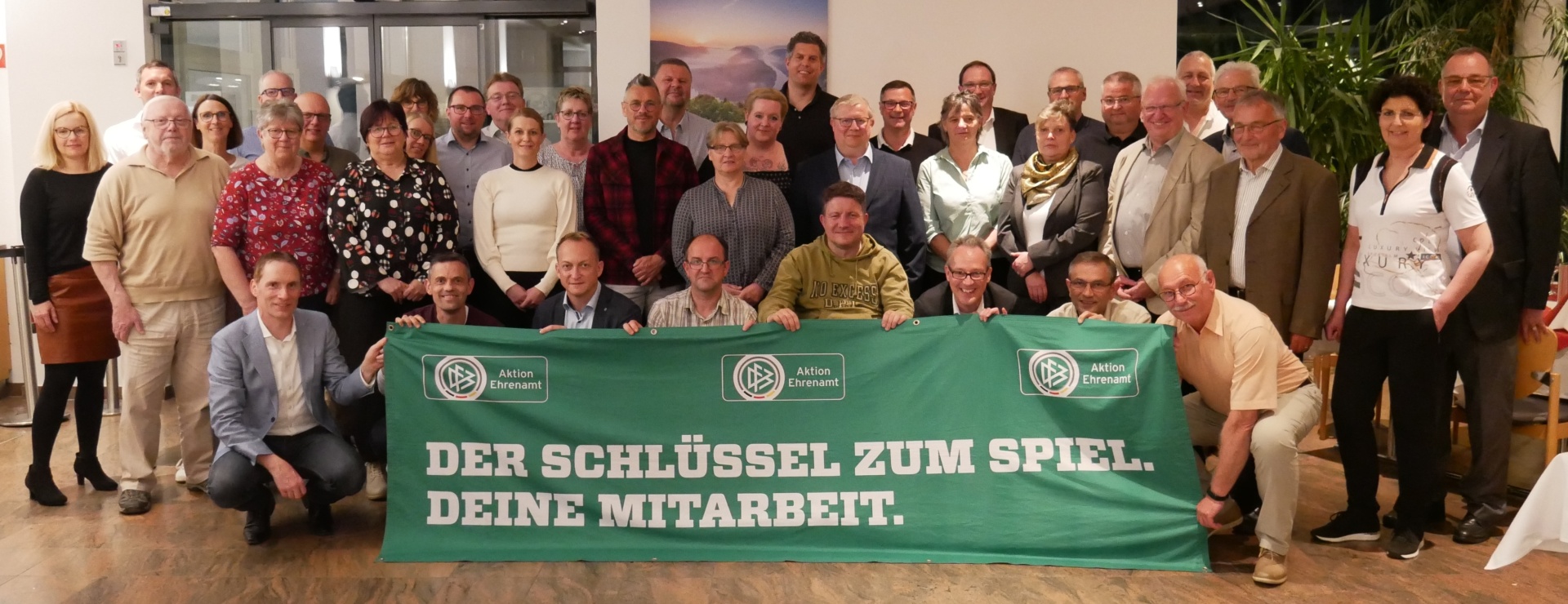 [{"clang_id":1,"value":"Gruppe von etwa 30 Personen vor grünem Banner mit Aufschrift \"DER SCHLÜSSEL ZUM SPIEL. DEINE MITARBEIT.\" im Innenraum."}]