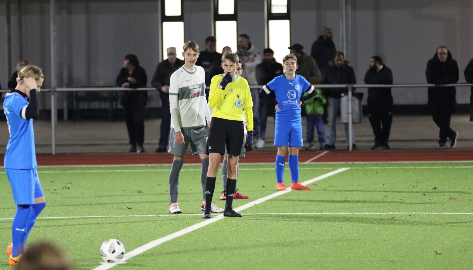 [{"clang_id":1,"value":"Fußballspiel im Abendlicht. Vier Spieler und Schiedsrichter stehen am Spielfeld, um den Ball. Im Hintergrund Zuschauer und Gebäude."}]