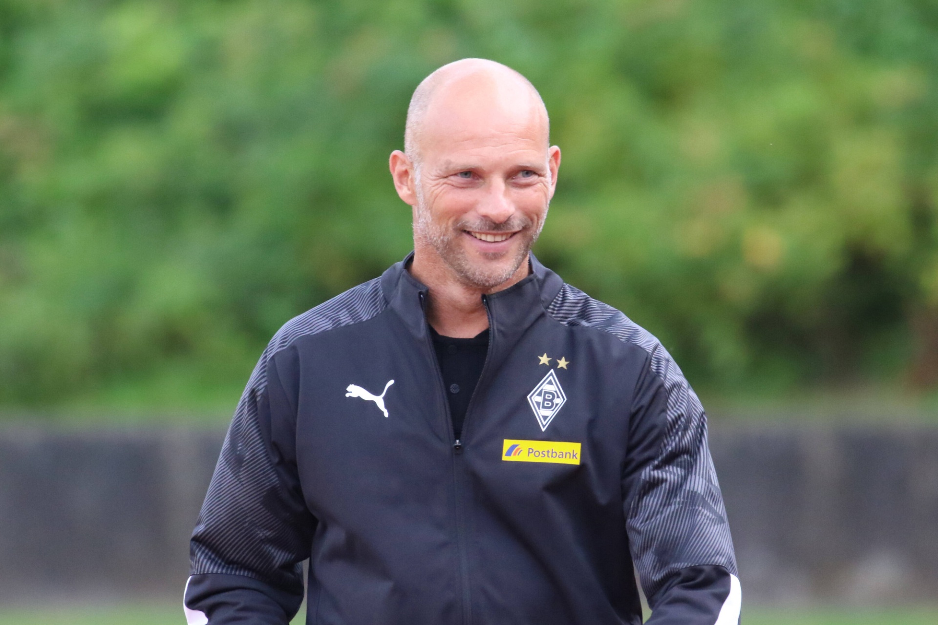 [{"clang_id":1,"value":"Mann mit Glatze und Bart lächelt in die Kamera. Er trägt eine schwarze Trainingsjacke mit Borussia Mönchengladbach Logo und PUMA-Schriftzug. Hintergrund ist Grün."}]